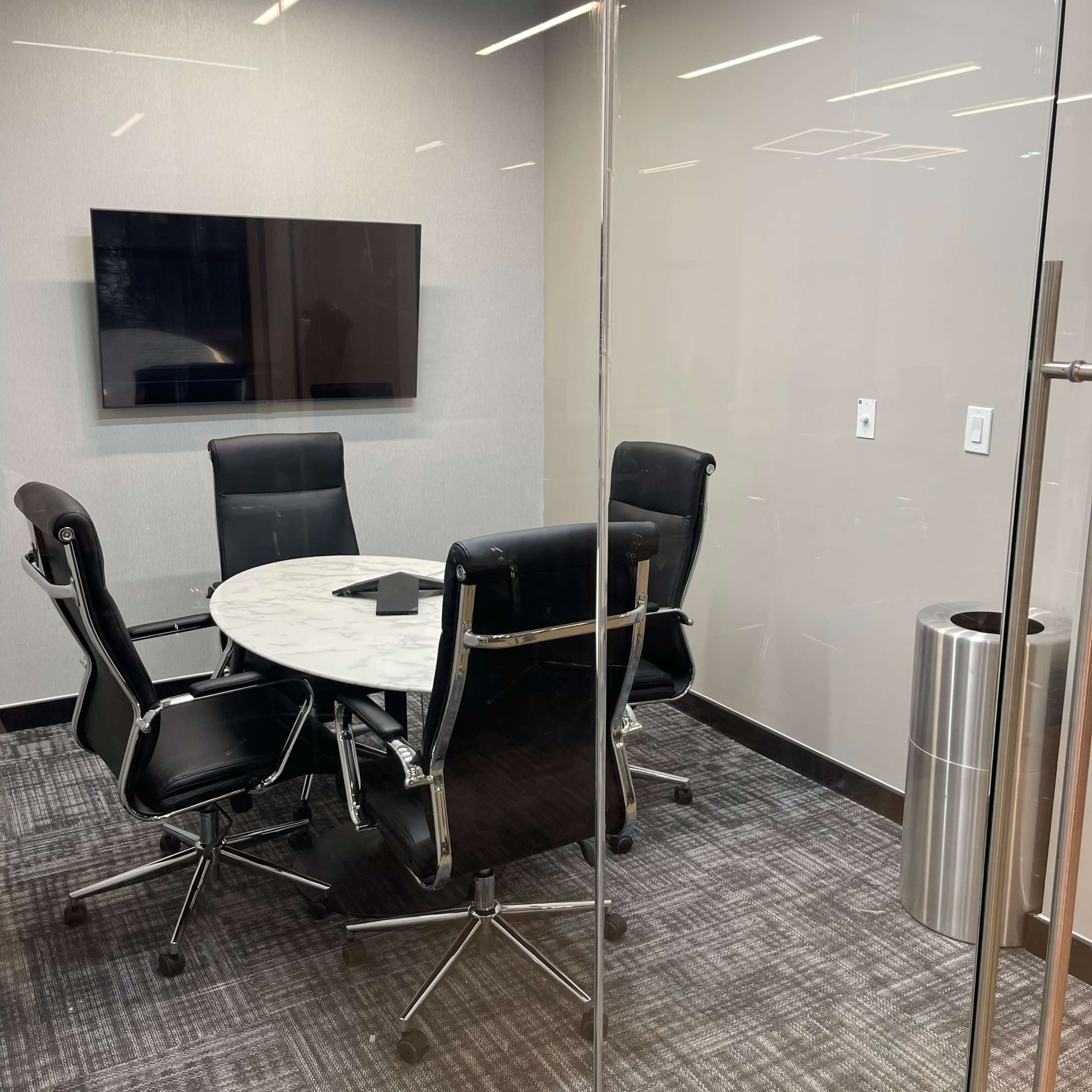 A glass-walled conference room features a round table surrounded by four black leather chairs, with a television mounted on the wall.