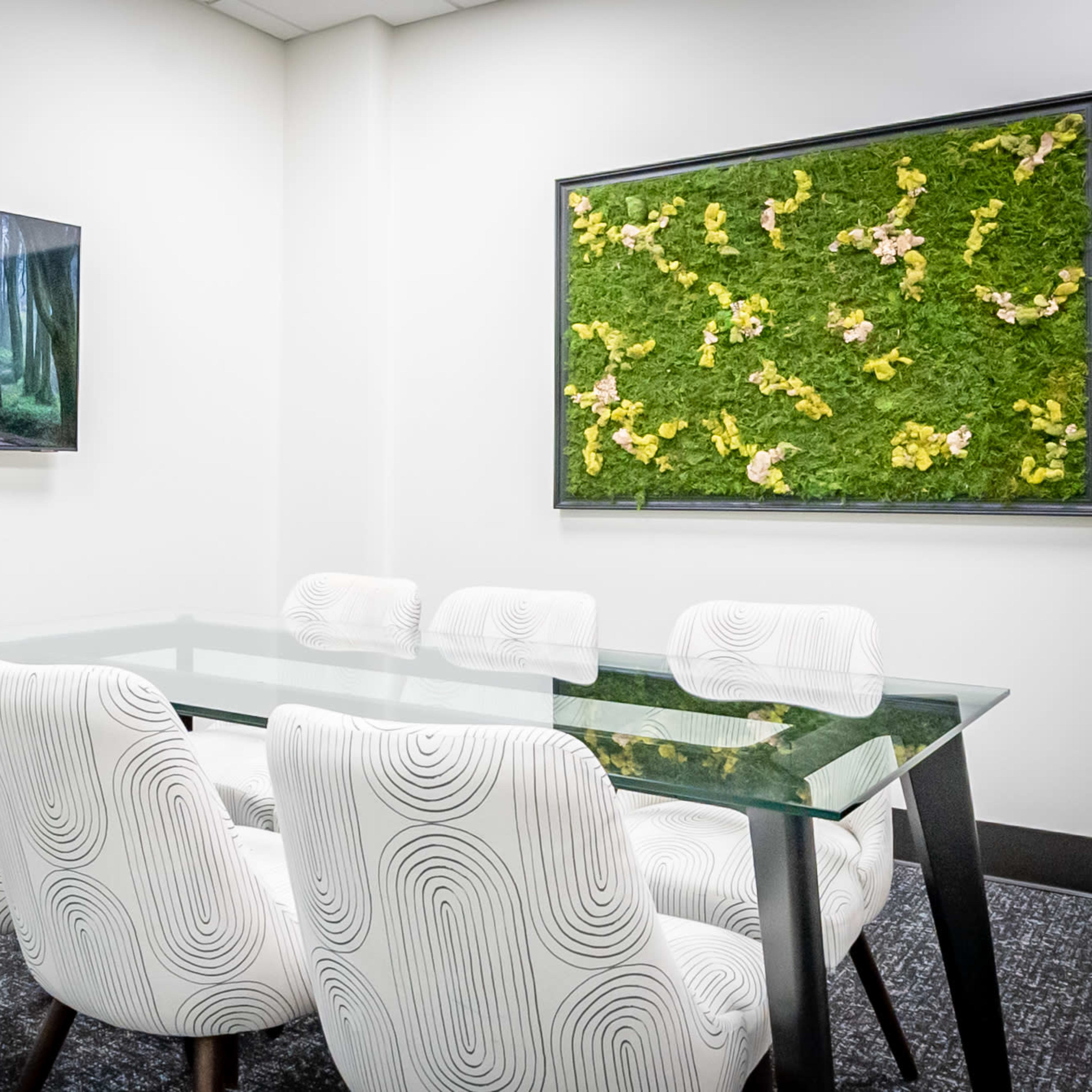 A modern conference room features a glass table surrounded by six white chairs, with a nature-themed artwork and a forest scene displayed on the walls.