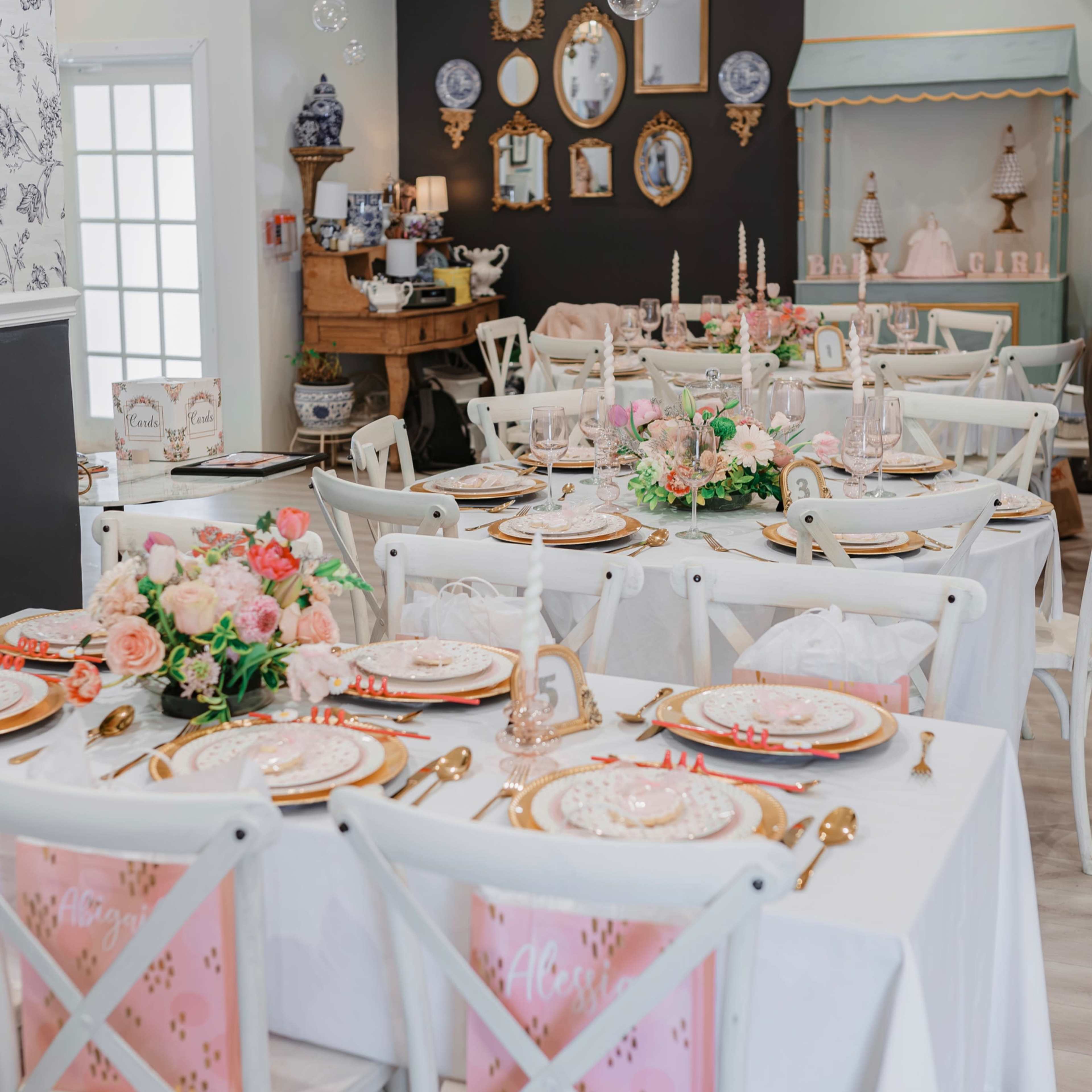 A dining area is set with white tables draped in tablecloths, adorned with pink accents, floral centerpieces, and decorative plates, surrounded by white chairs and framed mirrors on the walls.