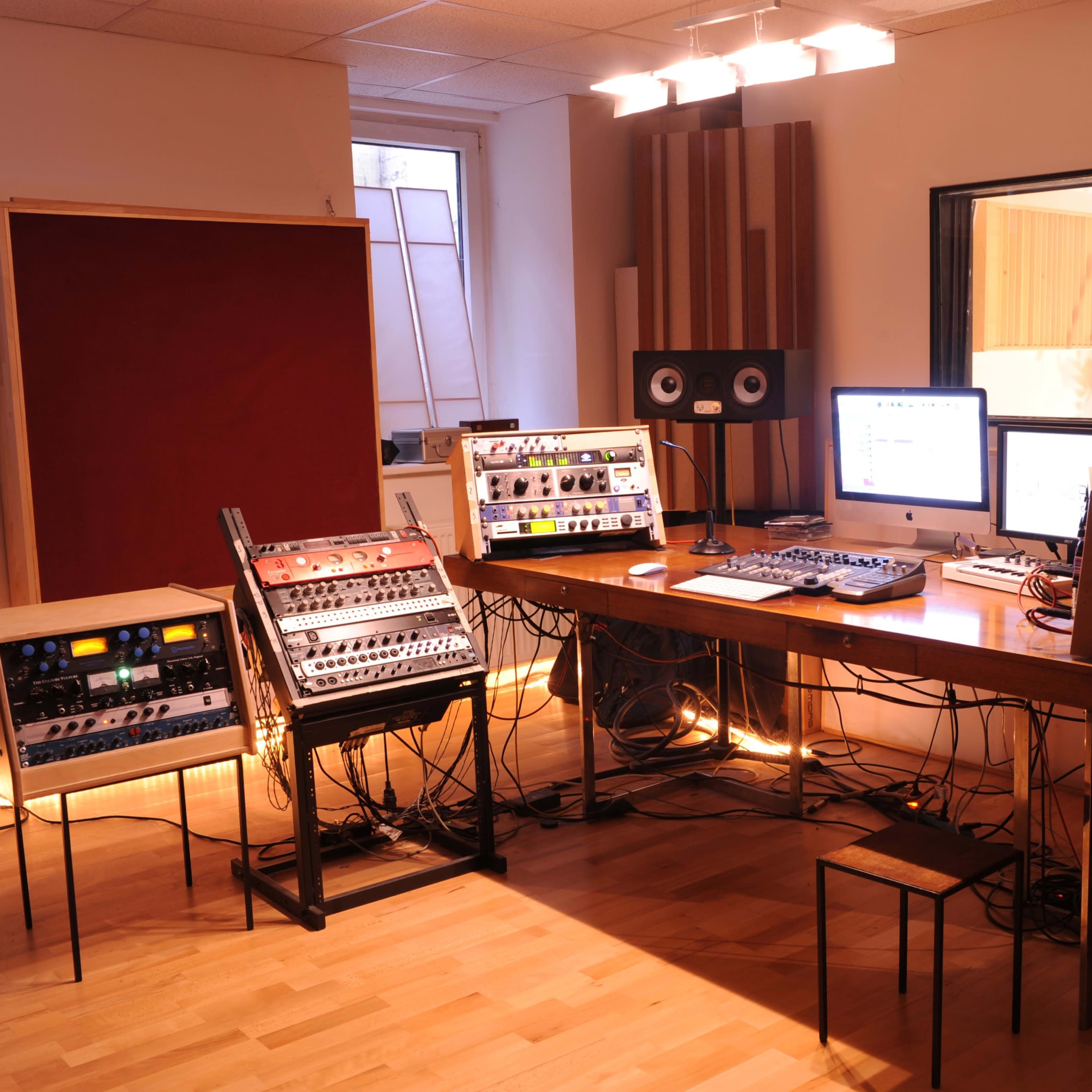 A music studio with audio equipment, a mixing desk, and monitors arranged around a wooden table in a well-lit room.