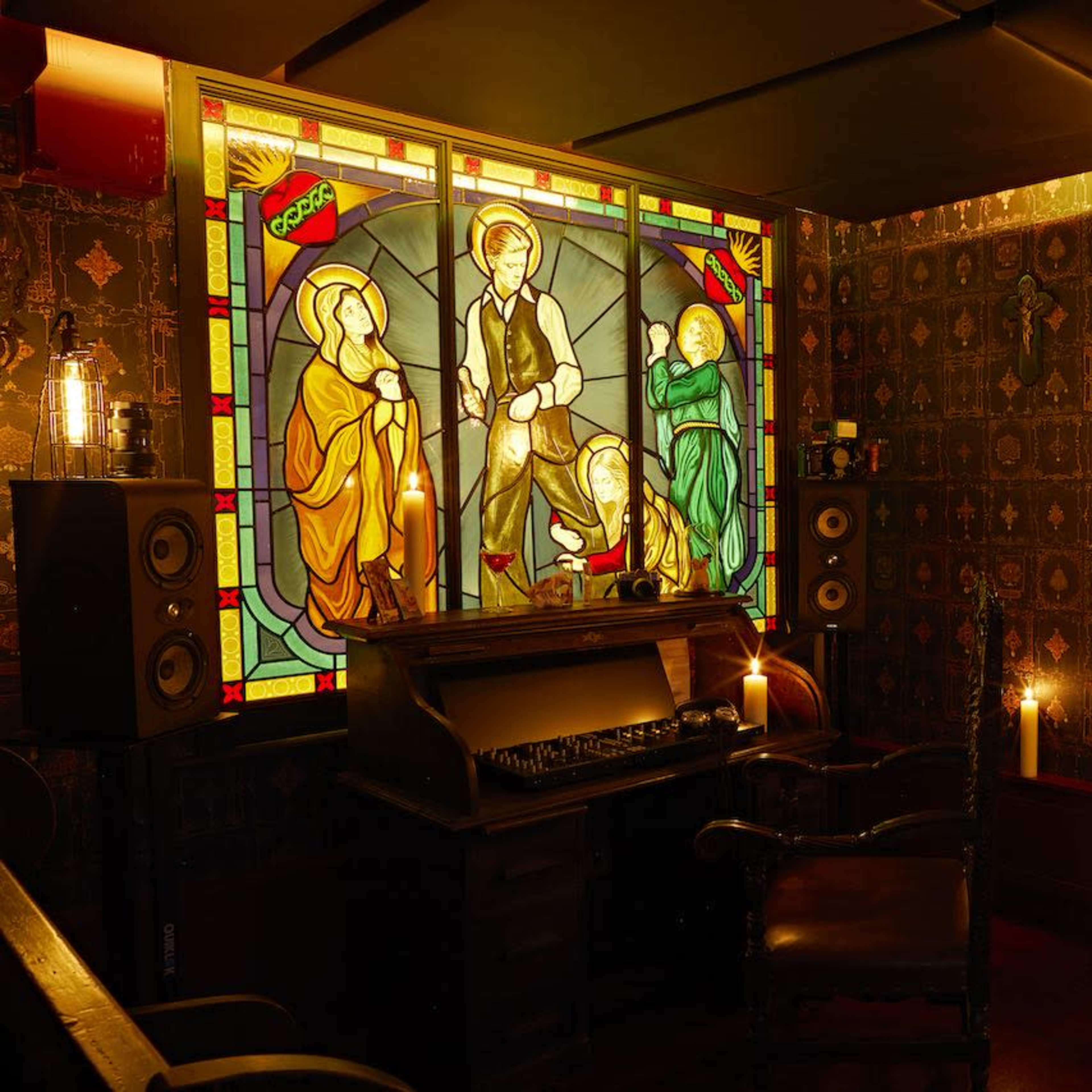 A dimly lit room with a stained glass window depicting three figures, an antique wooden desk with an old typewriter, a bookshelf filled with books, and various candles providing ambient light.