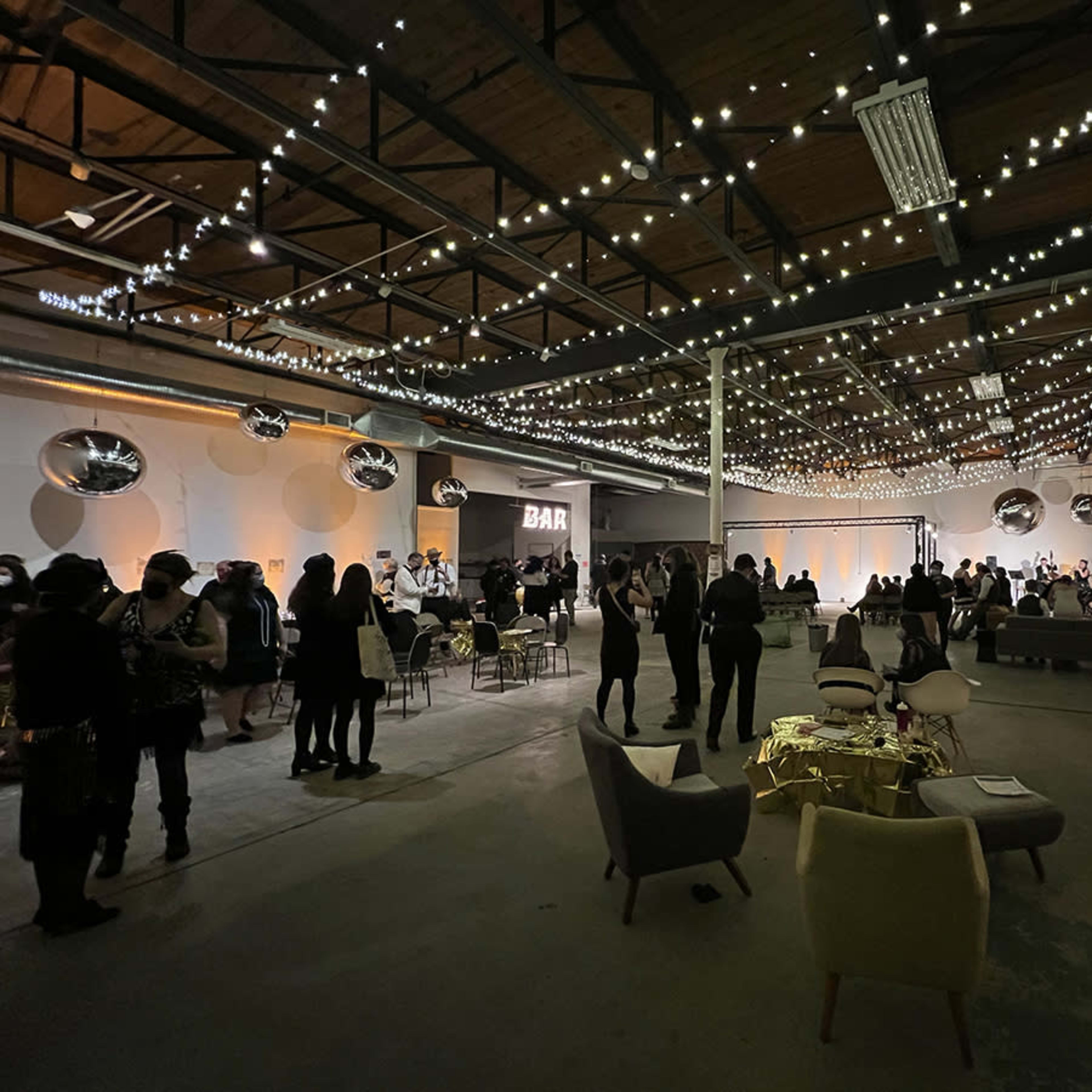 A spacious event venue decorated with string lights, featuring seated guests mingling and a bar area visible in the background.