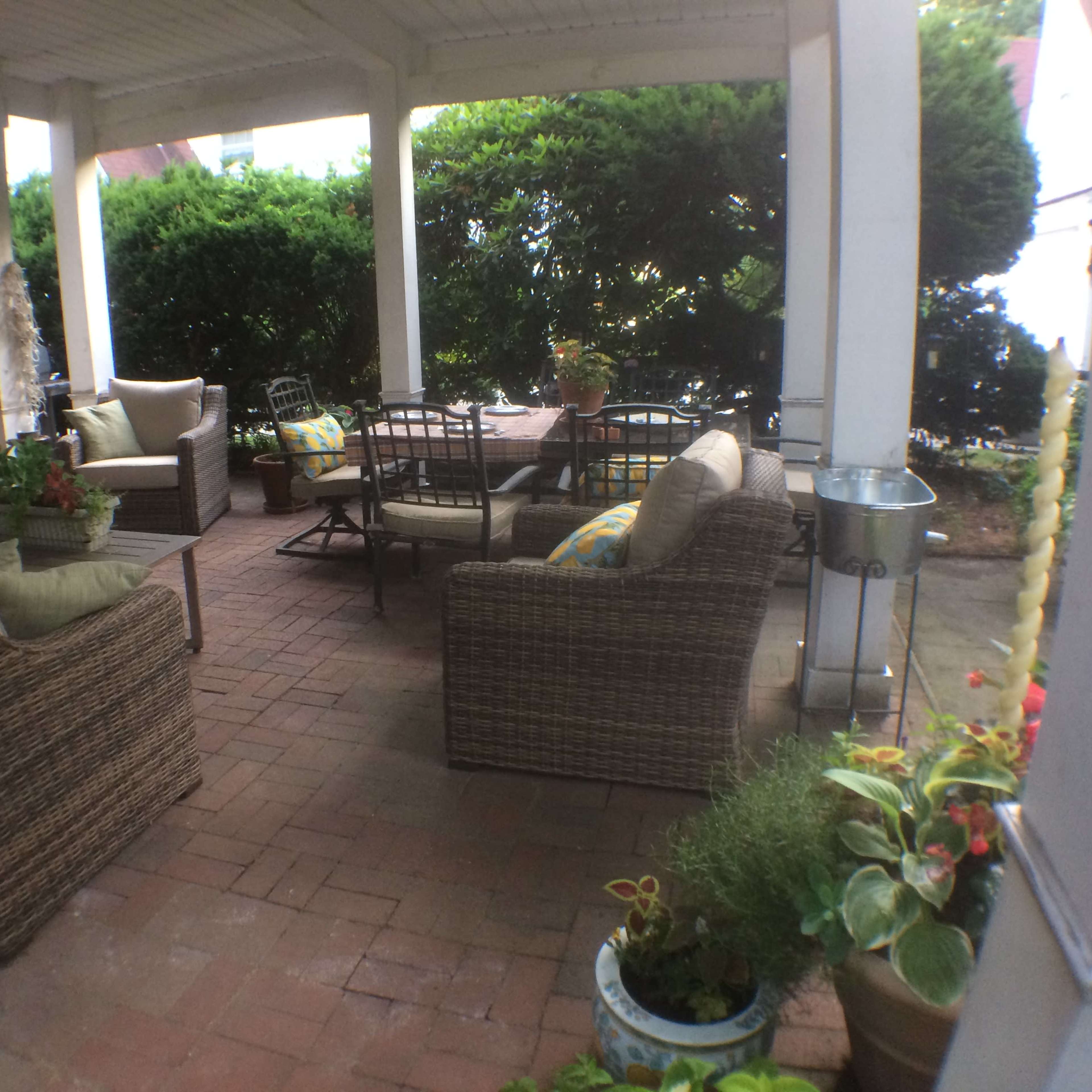 A covered outdoor patio area with wicker furniture, a dining table, and various potted plants.