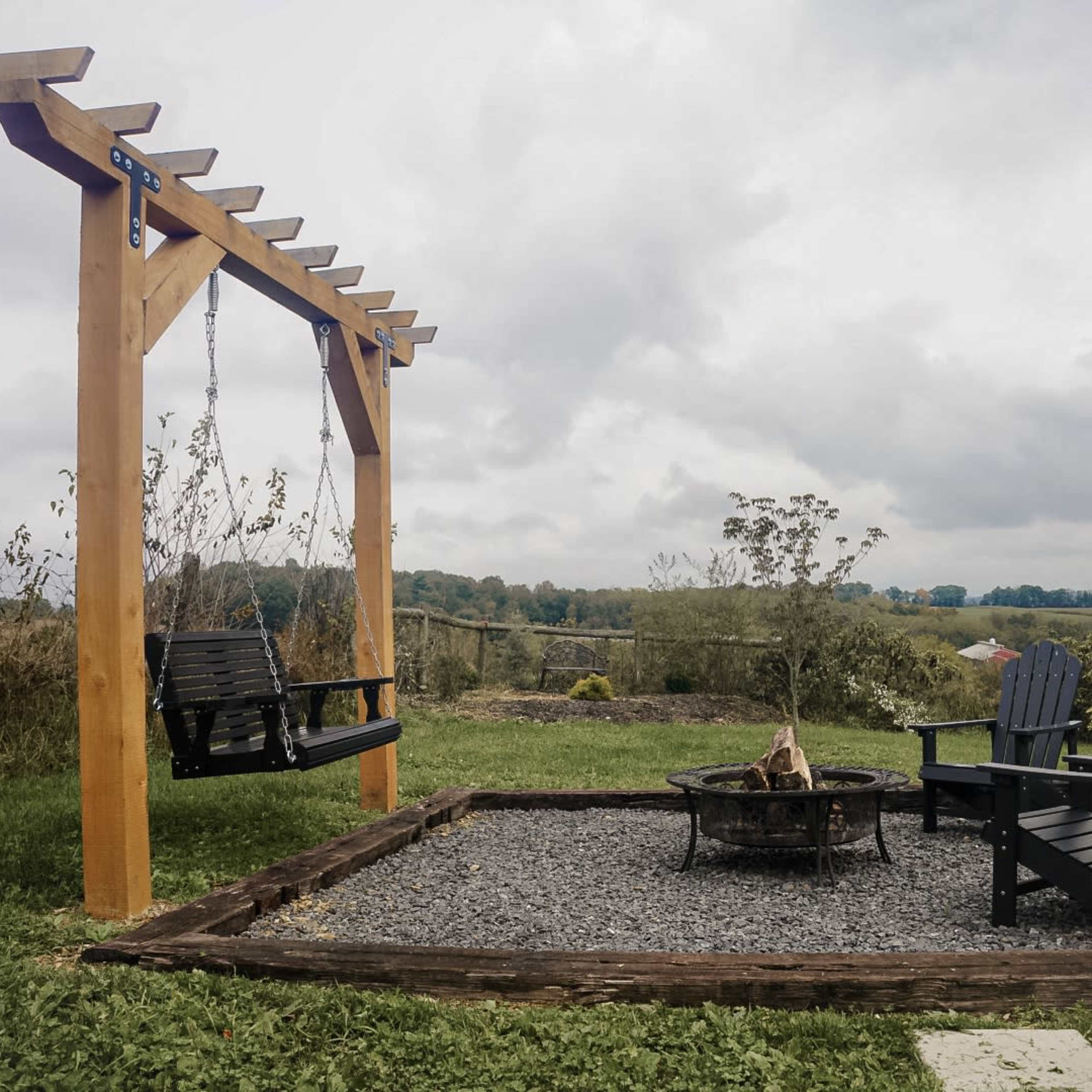 A wooden swing hangs from a pergola beside a fire pit, surrounded by two black chairs on a gravel base with rolling hills in the background.