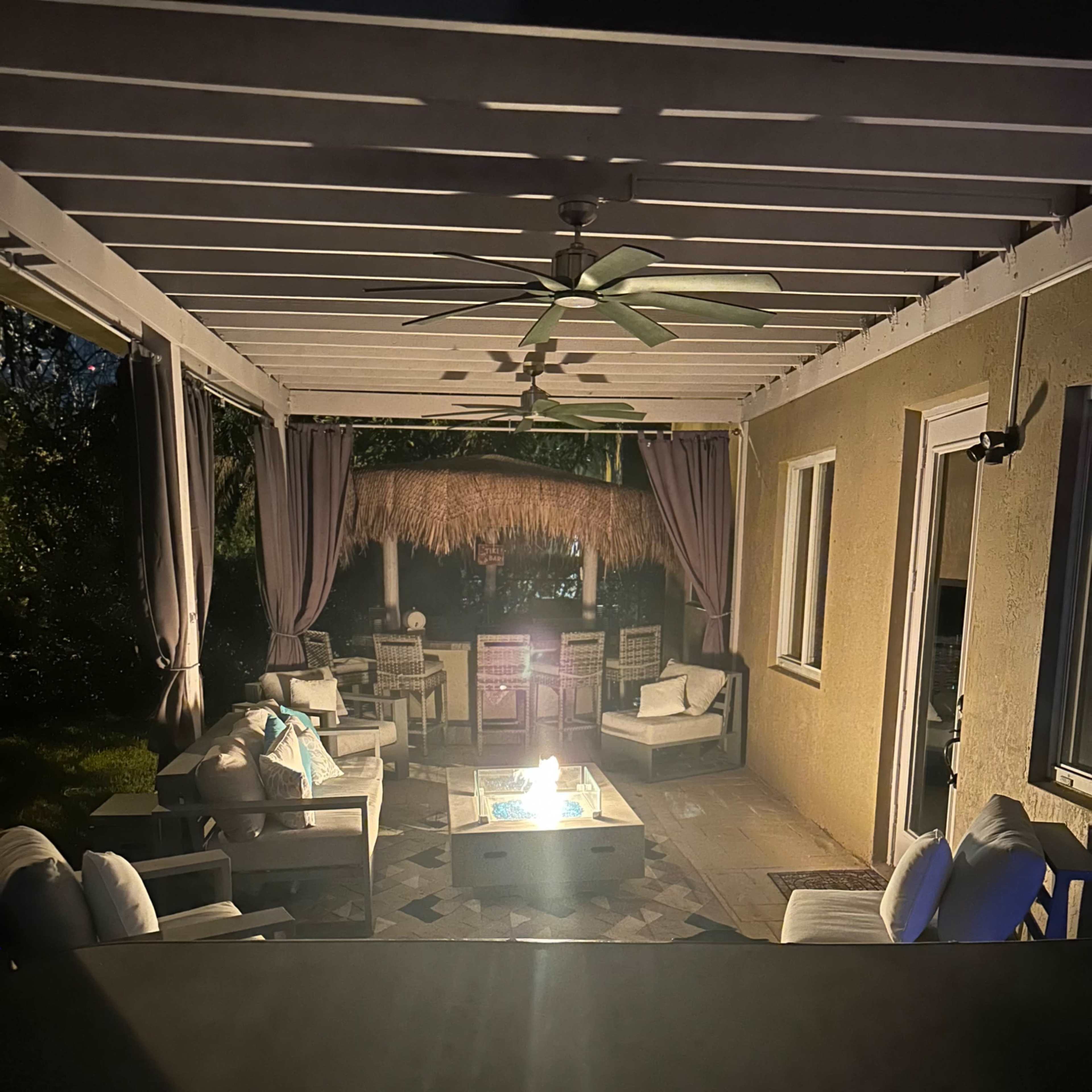 A cozy outdoor patio features a fire pit surrounded by seating, with a thatched awning overhead and soft lighting illuminating the scene.