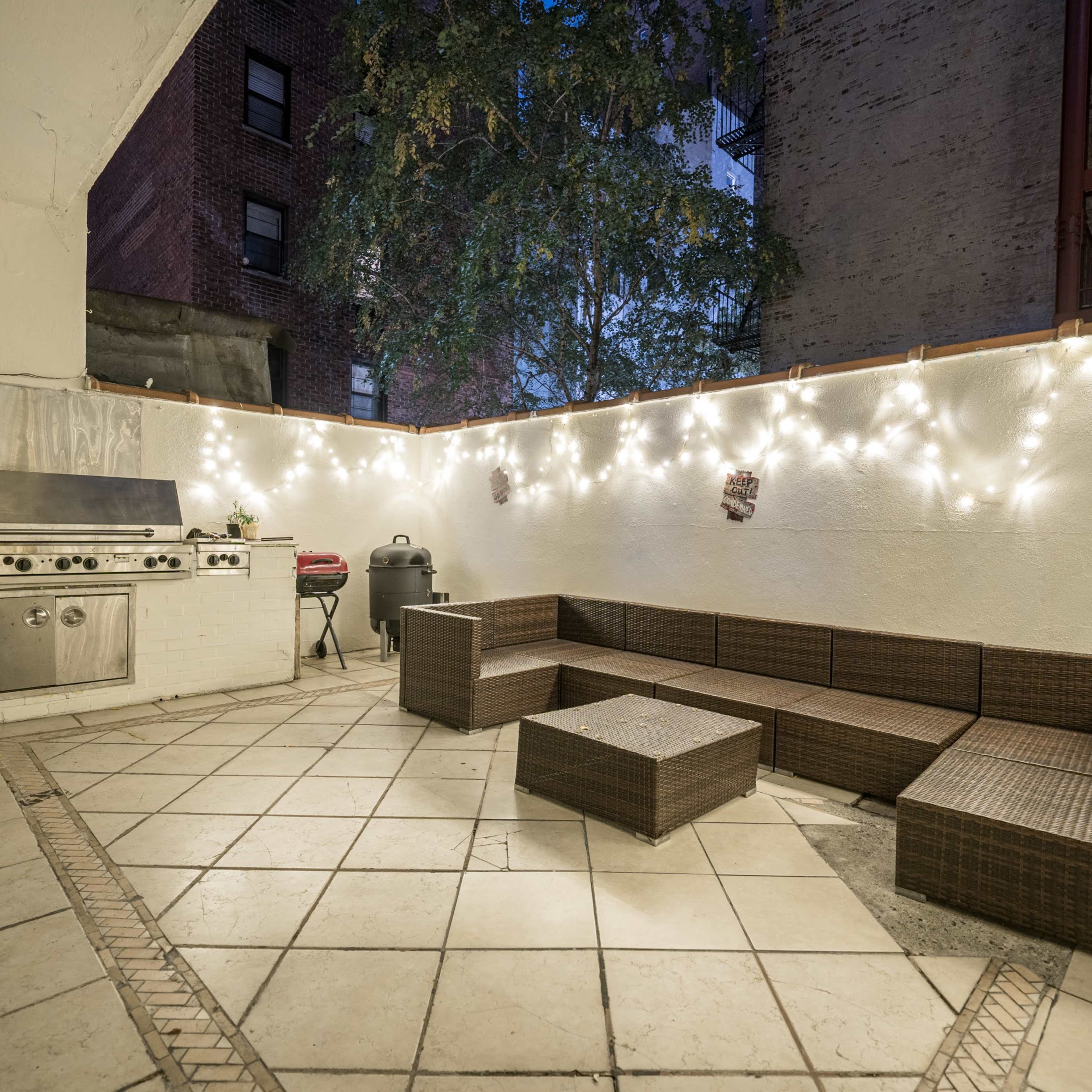 A patio area with a grill and a brown wicker seating arrangement illuminated by string lights at dusk.