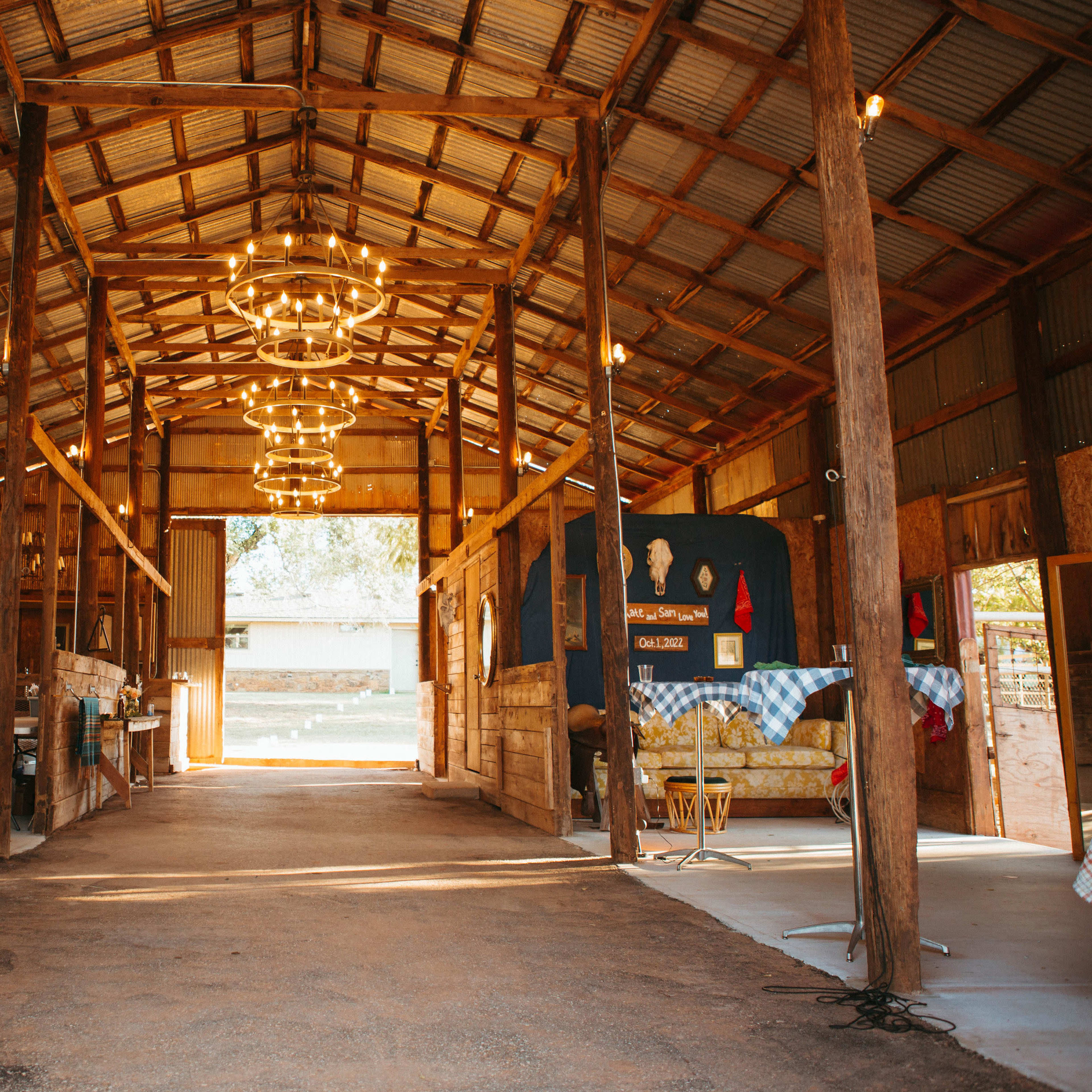 The interior of a large barn, featuring wooden beams, chandeliers, and tables set along the sides.