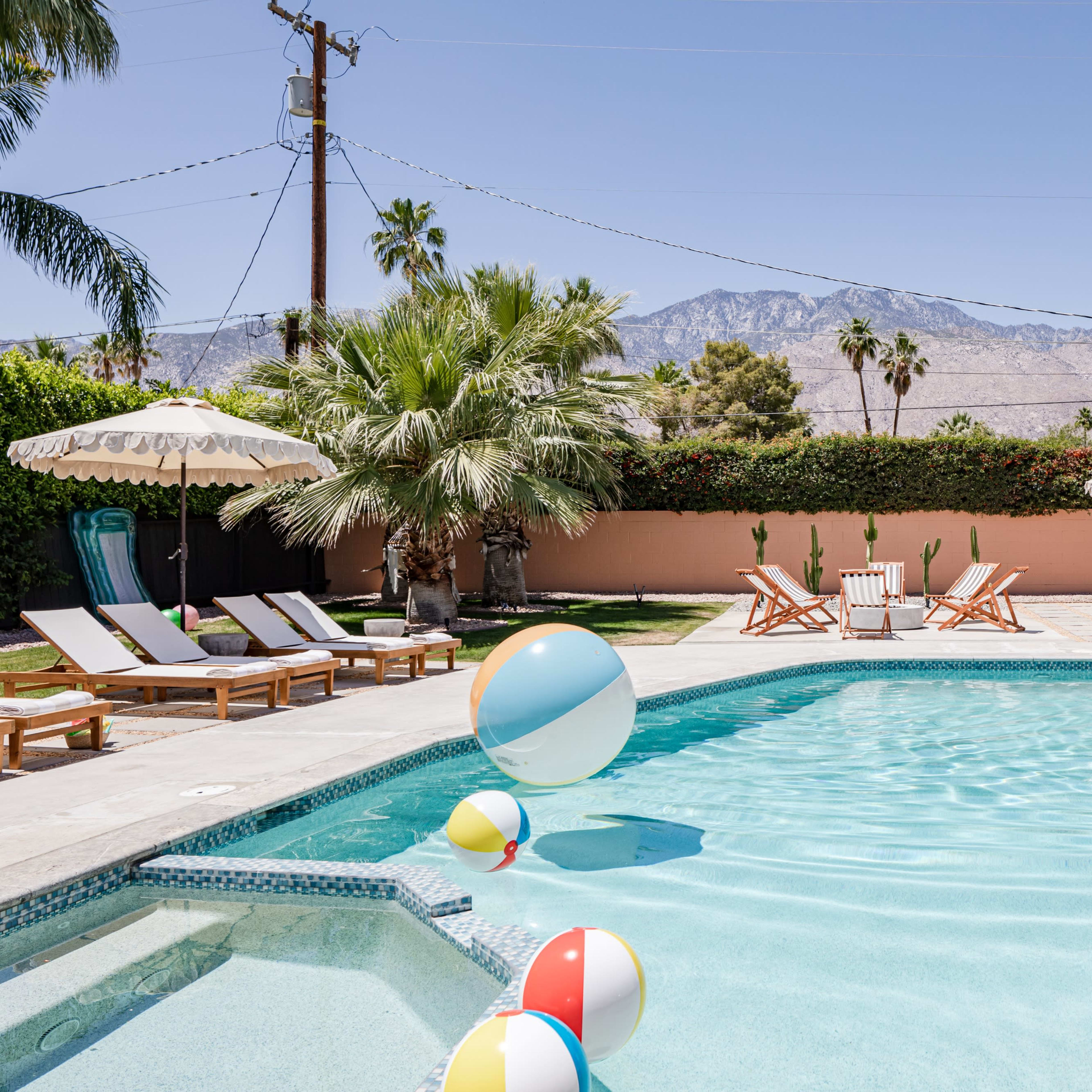 A sunny pool area features beach balls floating in the water, surrounded by lounge chairs, palm trees, and a backdrop of mountains.