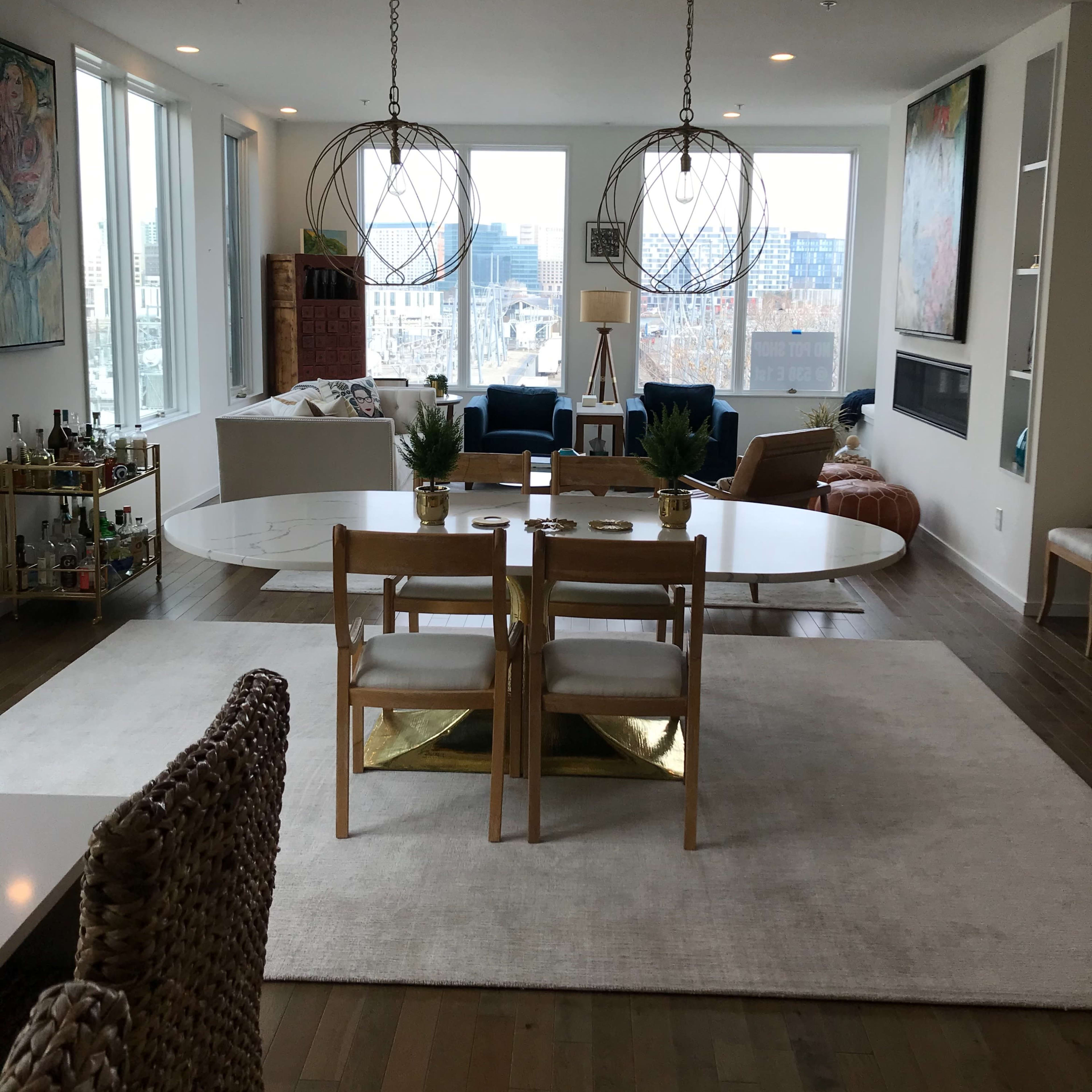 A modern living room features a large table surrounded by chairs, with large windows offering a view of the city and two spherical pendant lights hanging above.