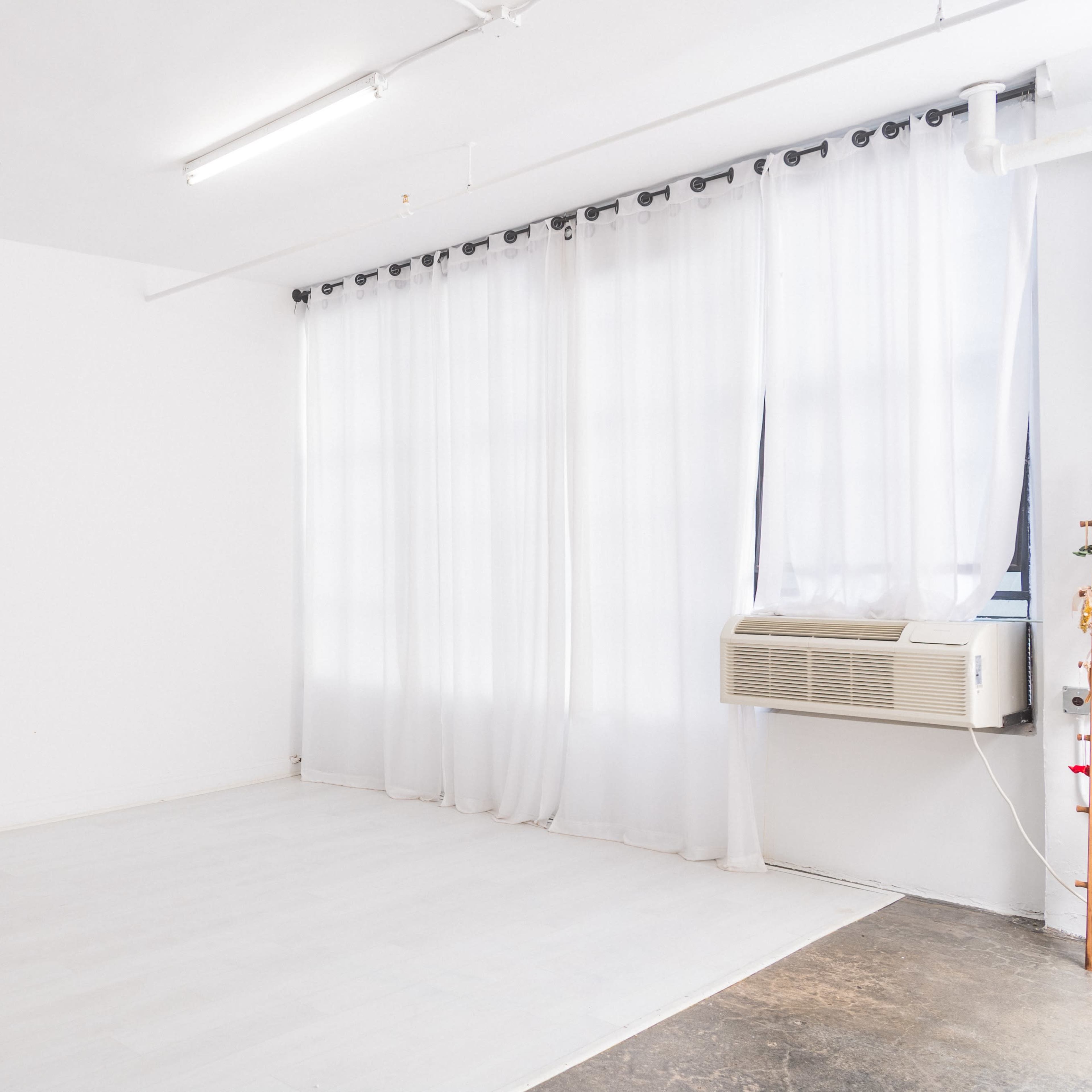 A spacious, minimally furnished room with white walls, a fan, large curtains, and an air conditioning unit.