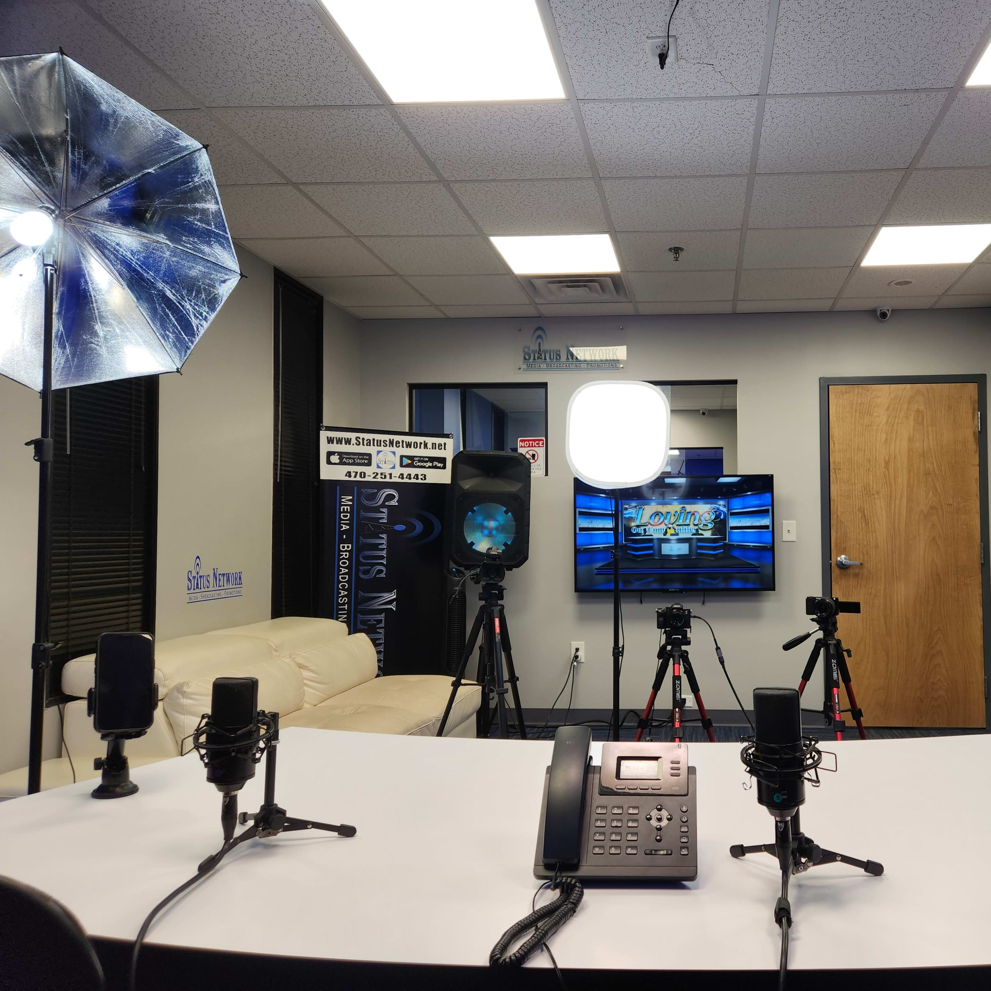 A video production studio with a table at the center, equipped with microphones and cameras, along with lighting setups and a monitor displaying graphics.