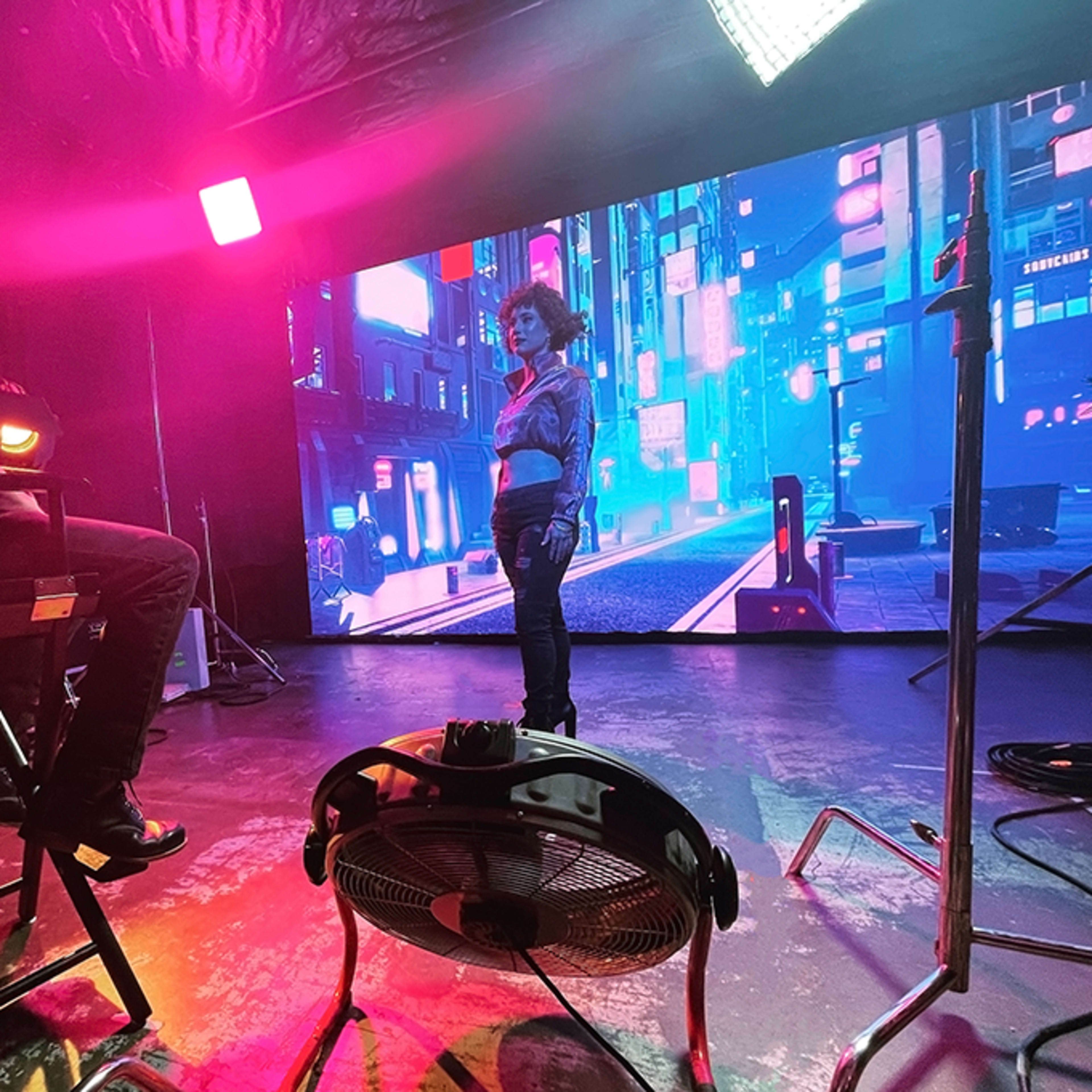 A person sits in a director's chair in front of a large screen displaying a vibrant cityscape, while another person stands on a set illuminated by colorful lights.