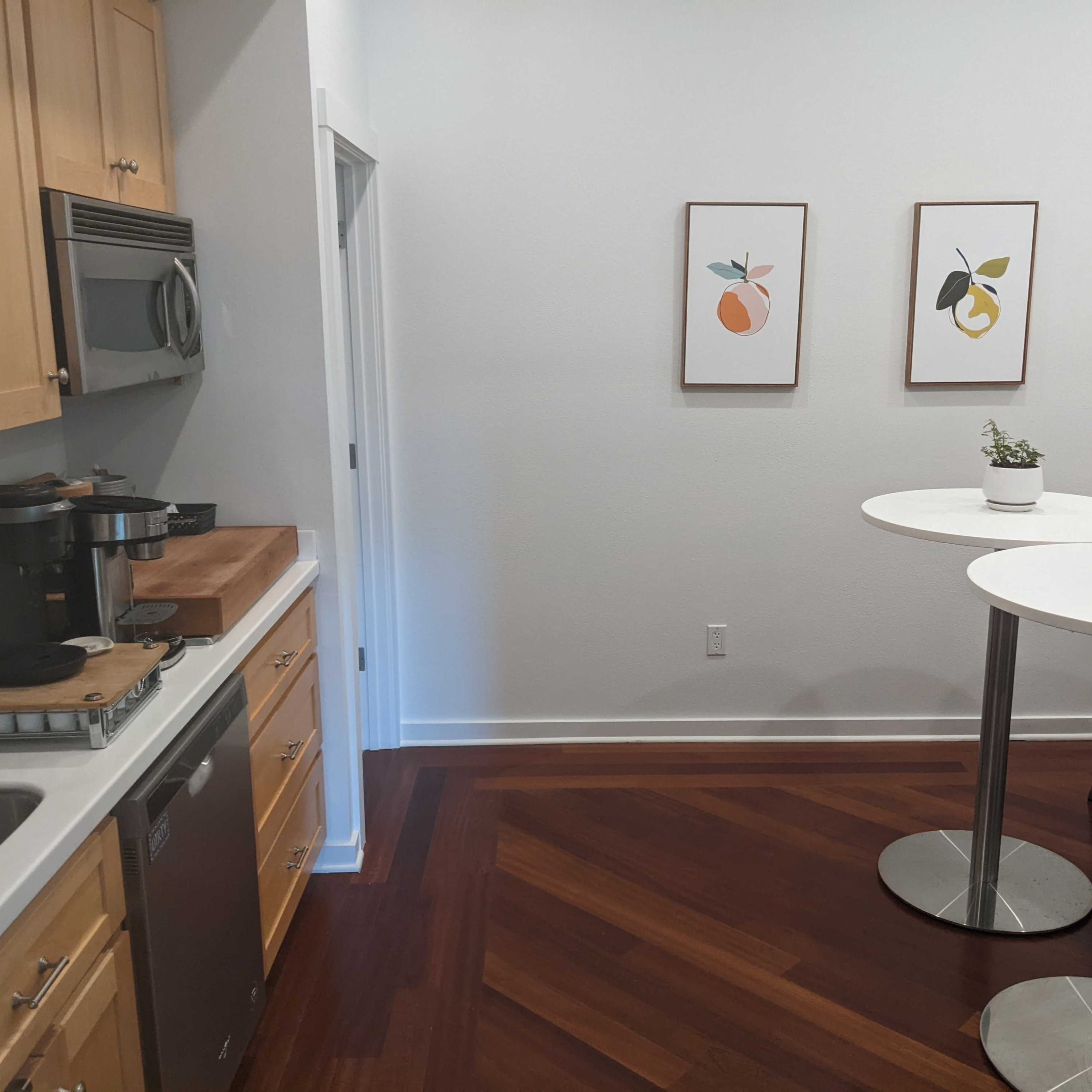 A modern kitchen area with wooden cabinets, a stainless steel sink, and two round white tables with metallic bases, adorned with framed fruit prints on the wall.