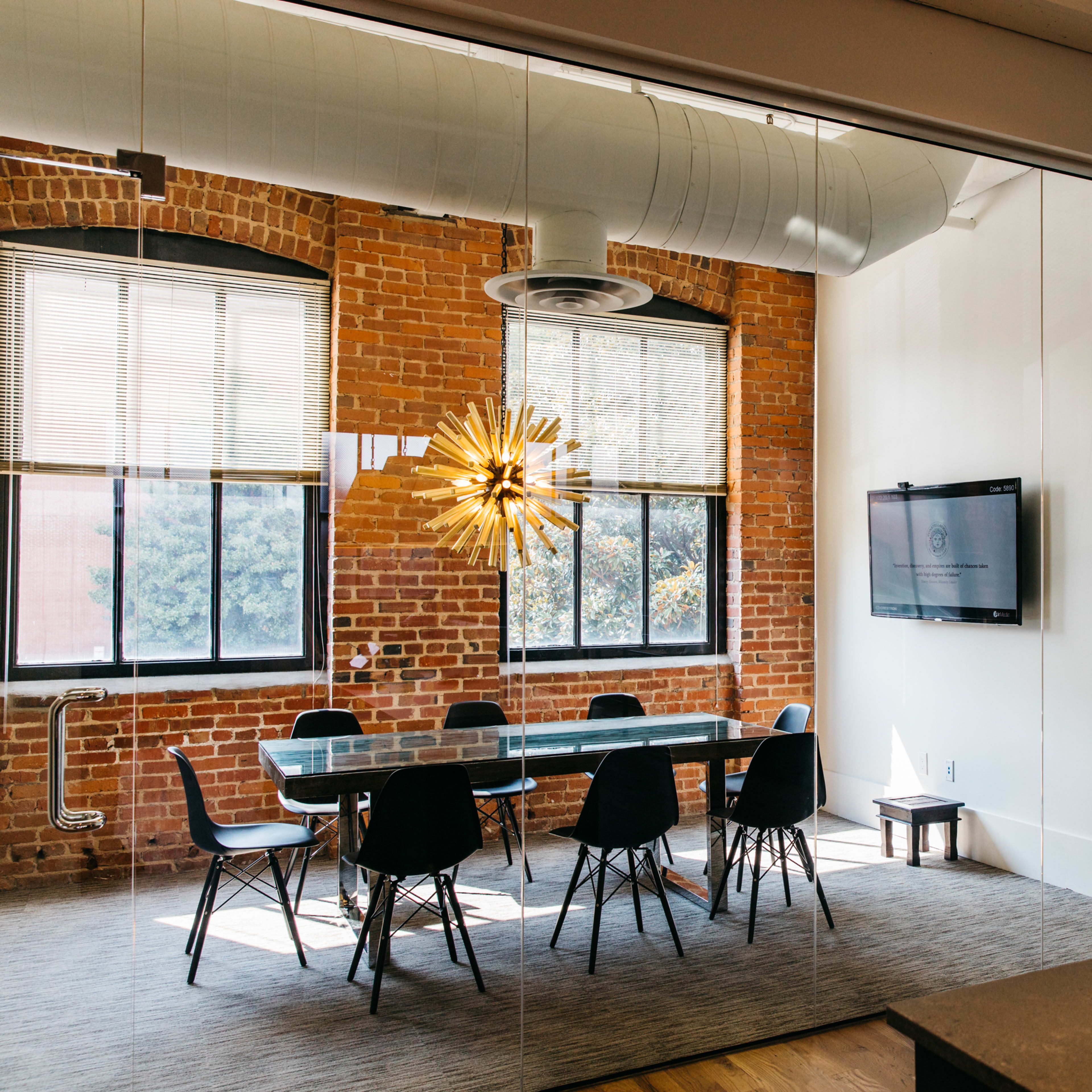 A bright conference room features a long table surrounded by black chairs, with large windows exposing brick walls and a television mounted on the wall.