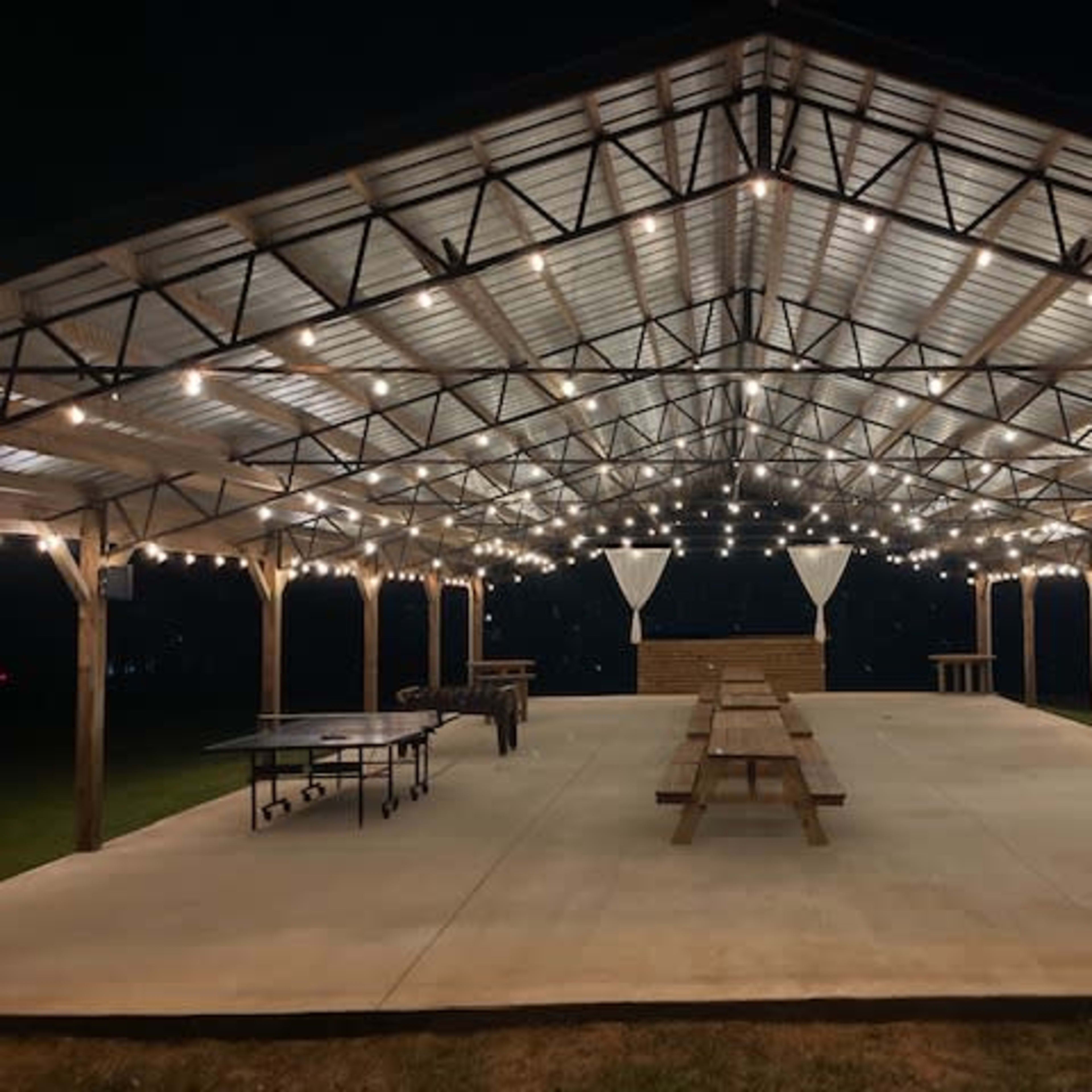 A large, open pavilion with string lights hanging from the ceiling, featuring multiple picnic tables and a concrete floor, set in a grassy area at night.