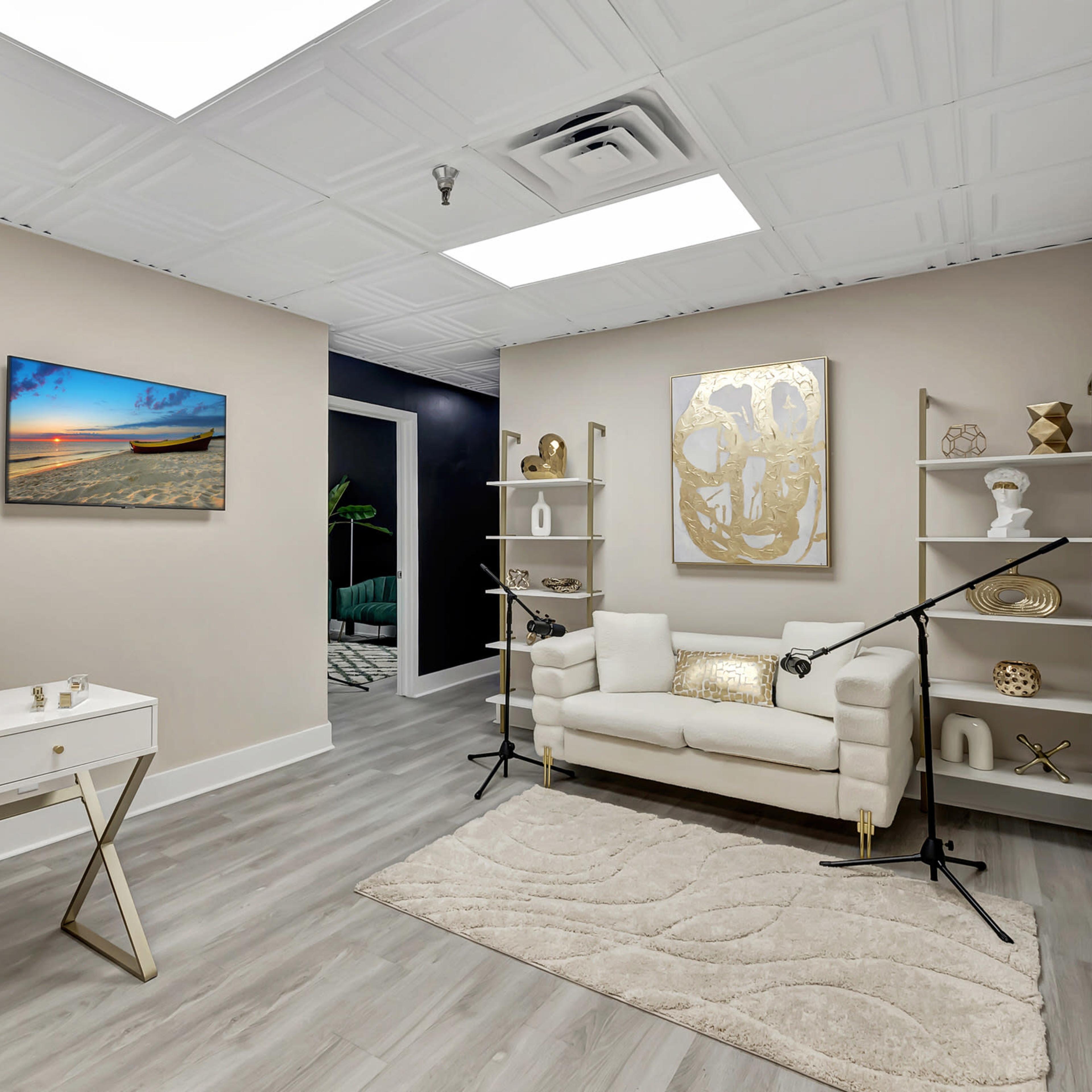 A modern office space featuring a white desk with a chair, a light-colored couch, decorative shelves, and a beach scene painting on the wall.