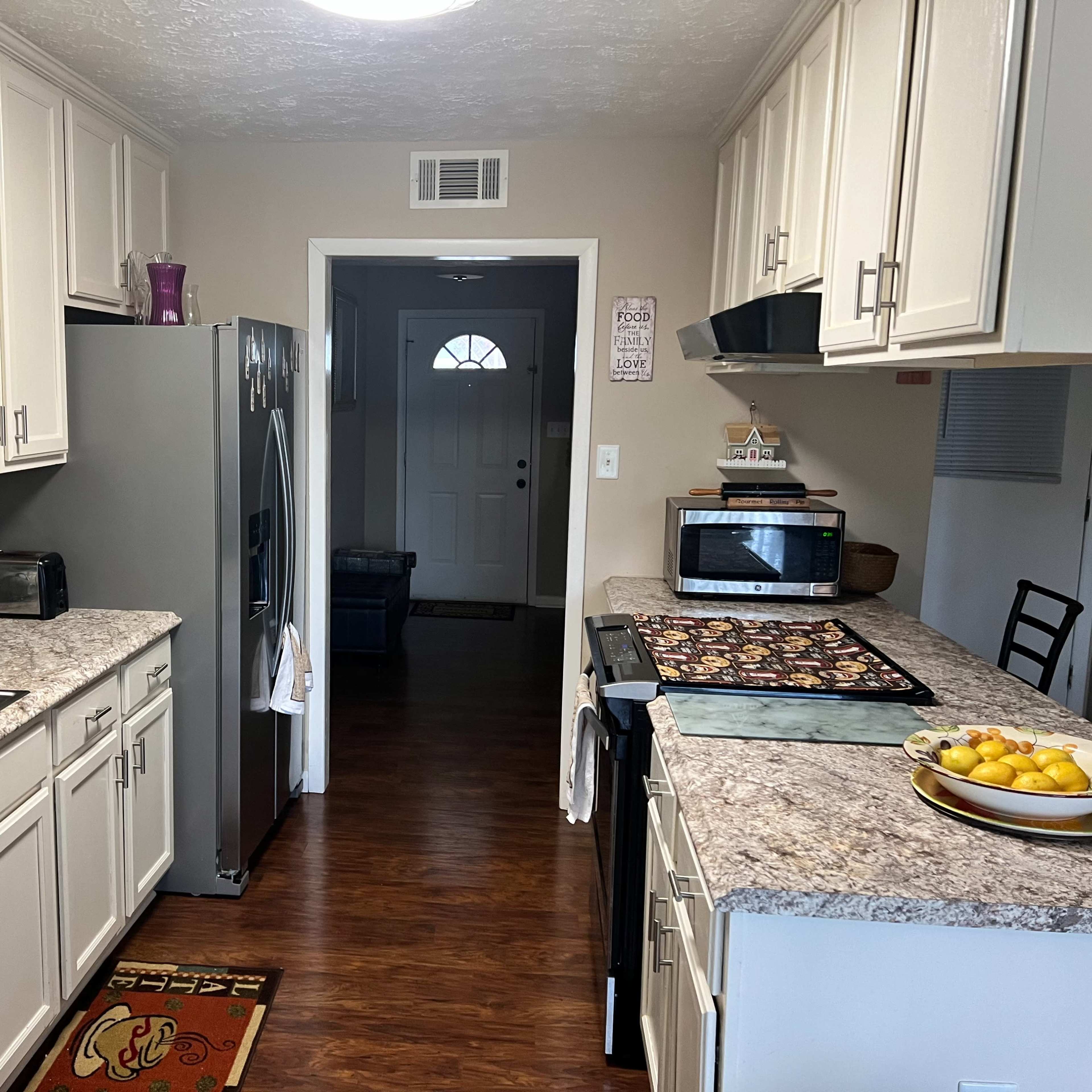A kitchen with light-colored cabinets, a granite countertop, and several appliances, including a refrigerator and microwave, with a view into a hallway leading to a door.
