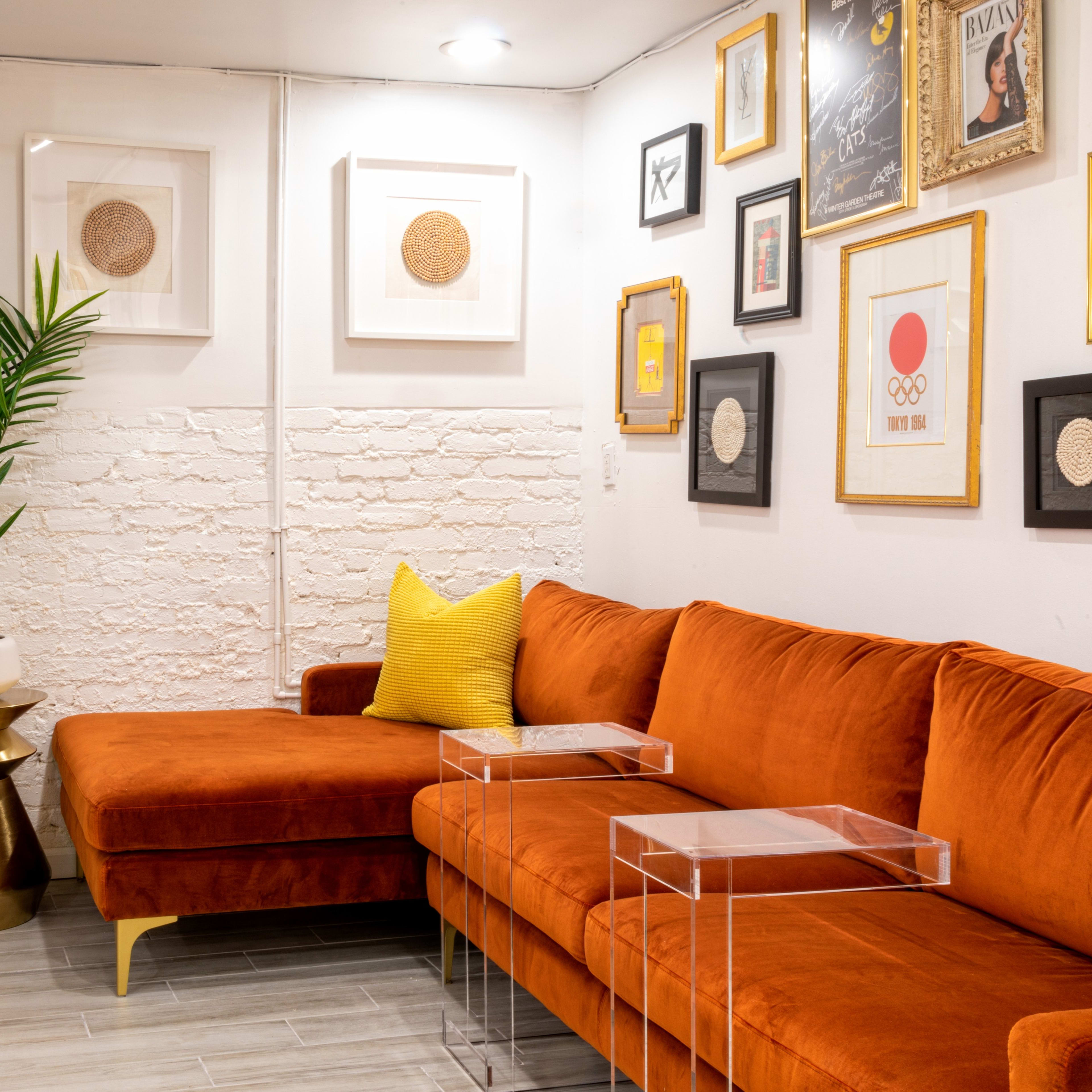 A cozy seating area with an orange sectional sofa, a small glass side table, a gold accent table, and framed artwork on the walls, complemented by a potted plant and curtains.