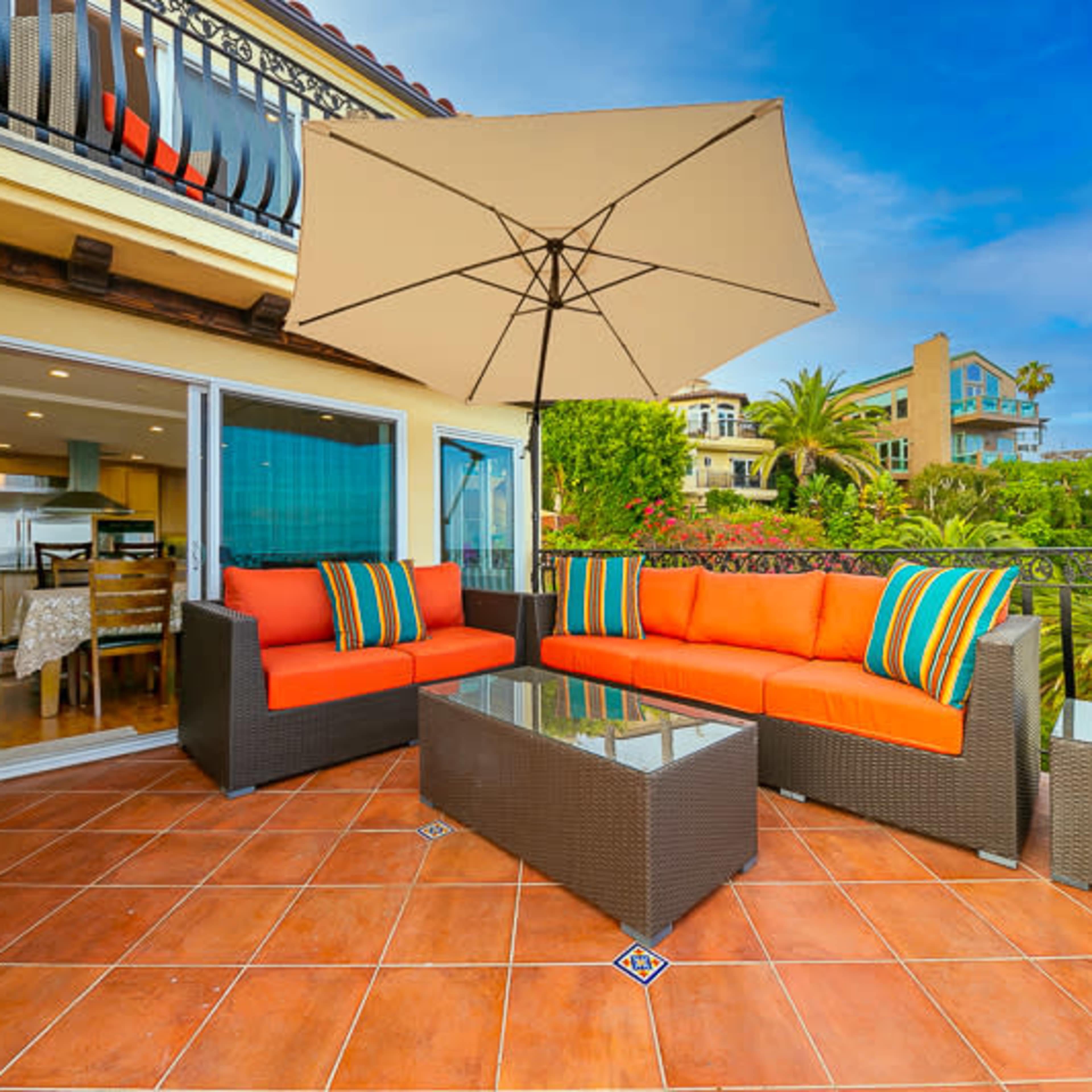 A patio area features orange cushions on wicker furniture under a large umbrella, overlooking a lush landscape and a residential backdrop.