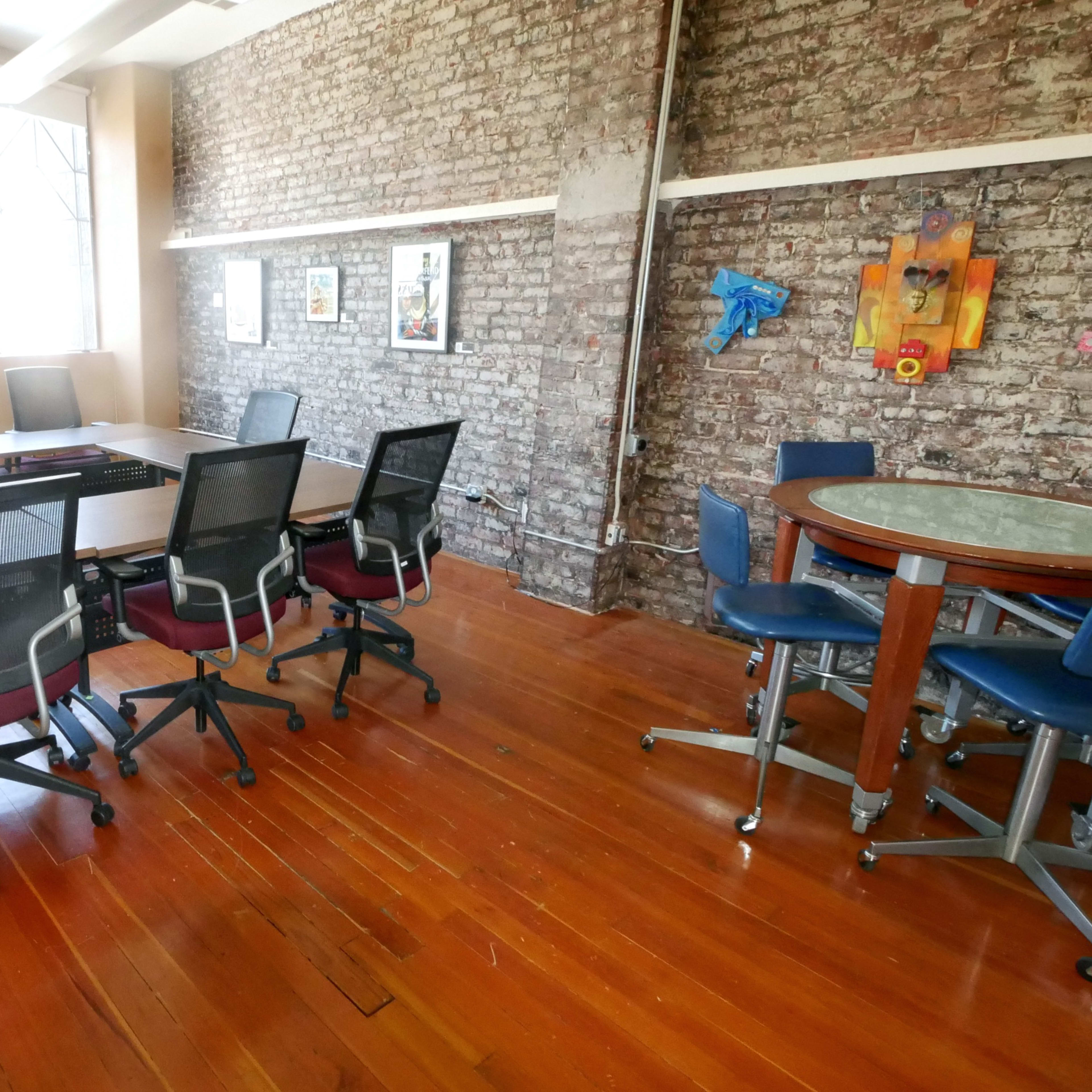 A conference room with a long table surrounded by chairs, large windows, and a brick wall.