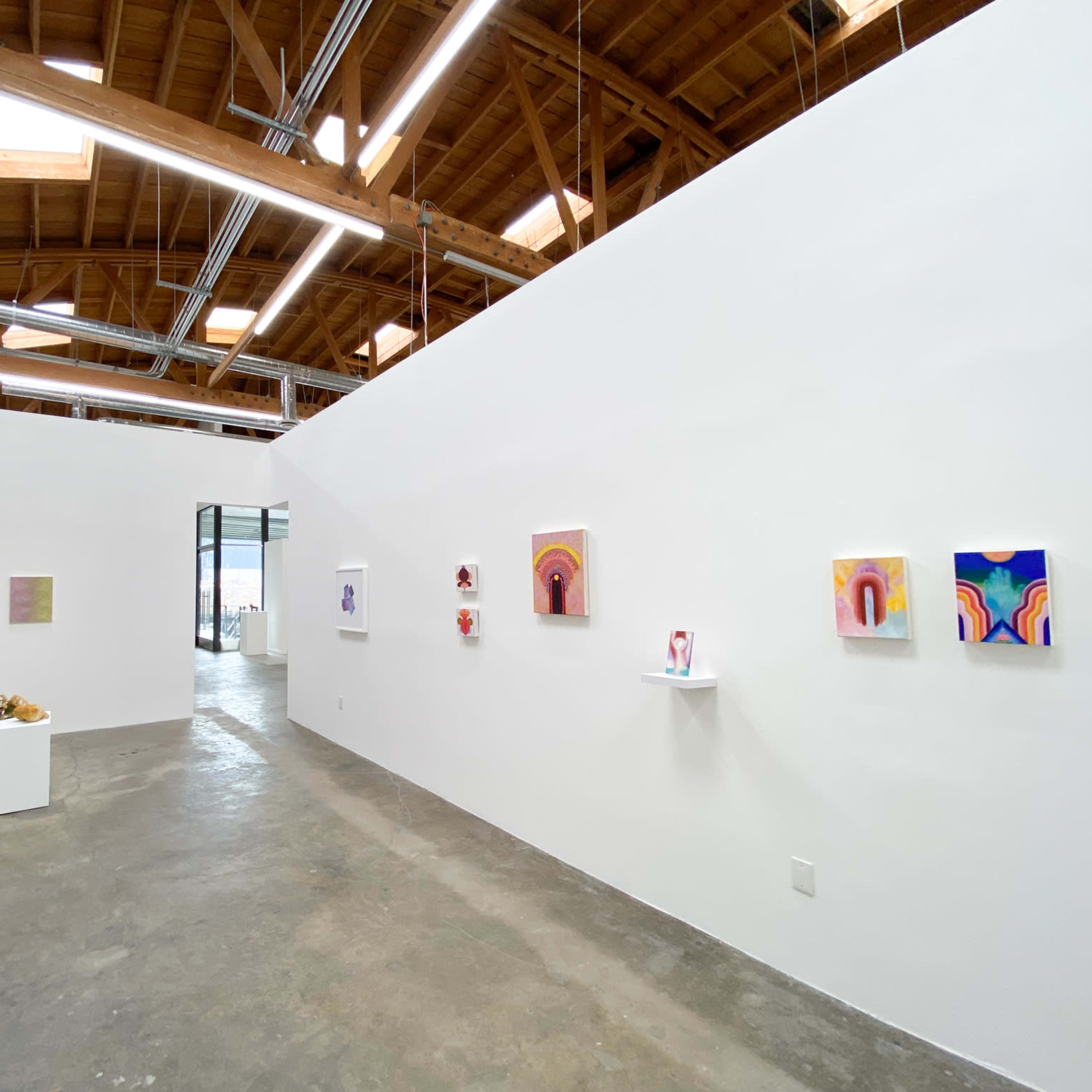 An art gallery with white walls featuring various paintings and small sculptures displayed on shelves and a table.