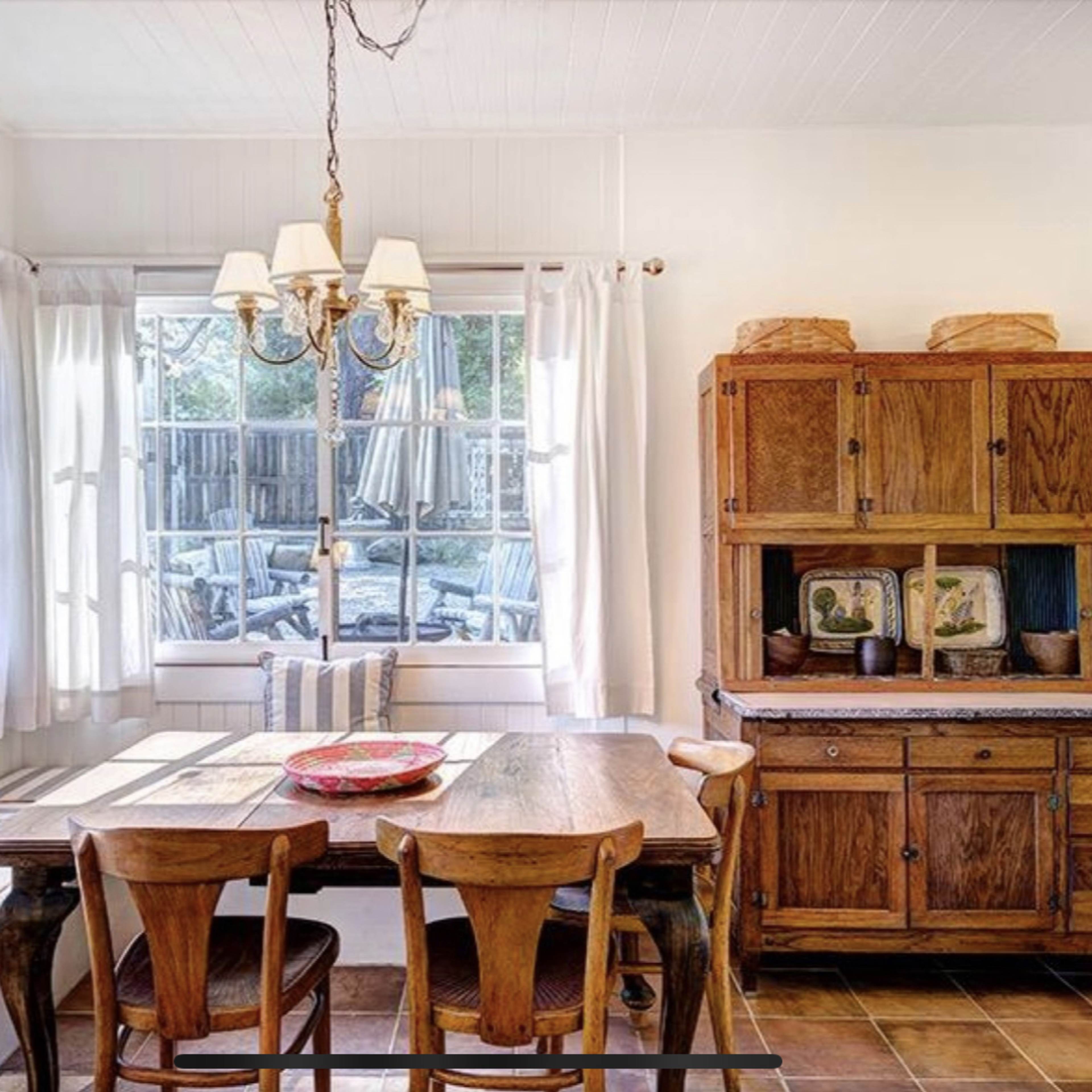 A wooden dining table with chairs is set in a bright kitchen, near large windows that overlook an outdoor space.