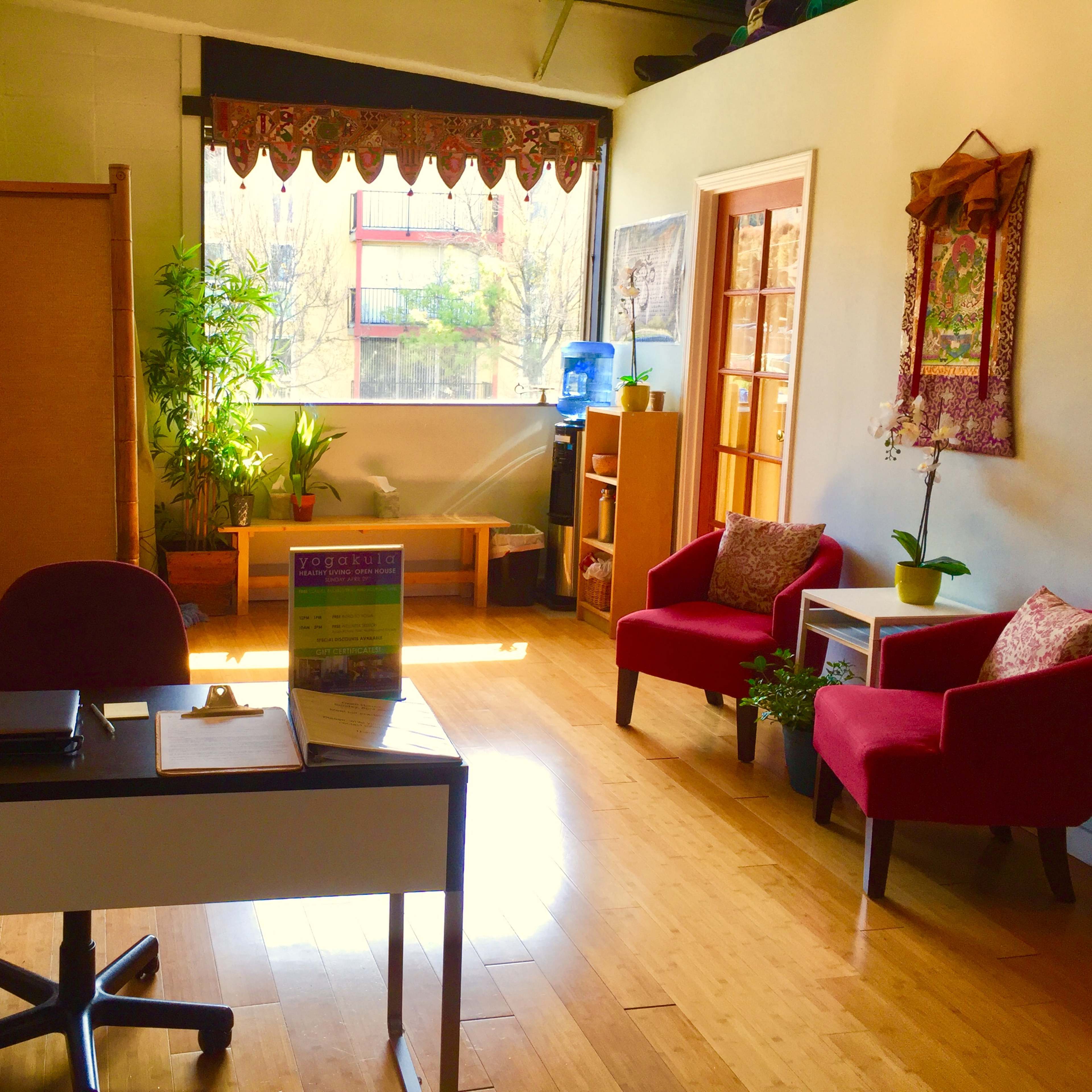 An office space with a desk, two red chairs, plants, and large windows letting in natural light.