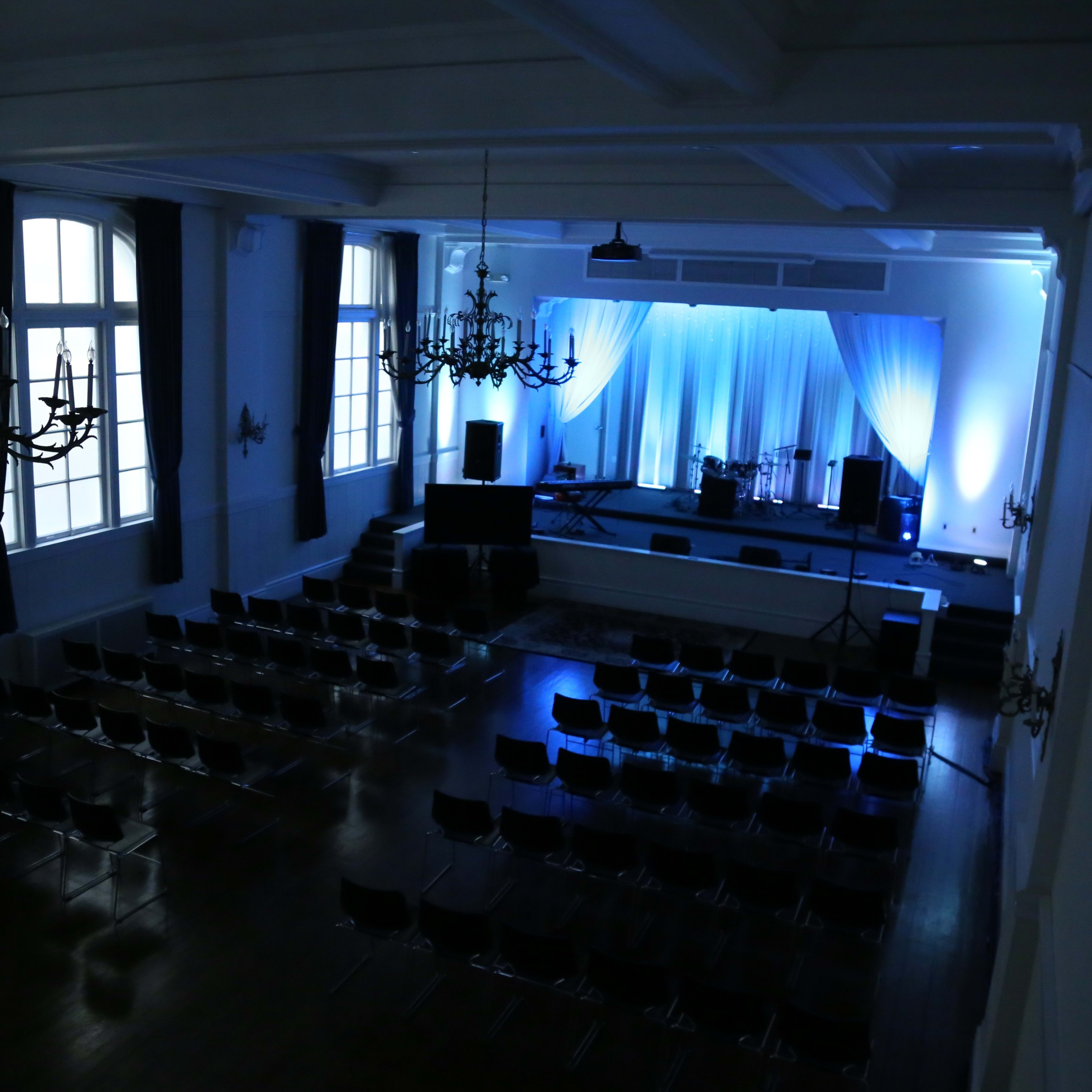 A spacious auditorium with rows of chairs facing a stage adorned with blue lighting and a white backdrop.
