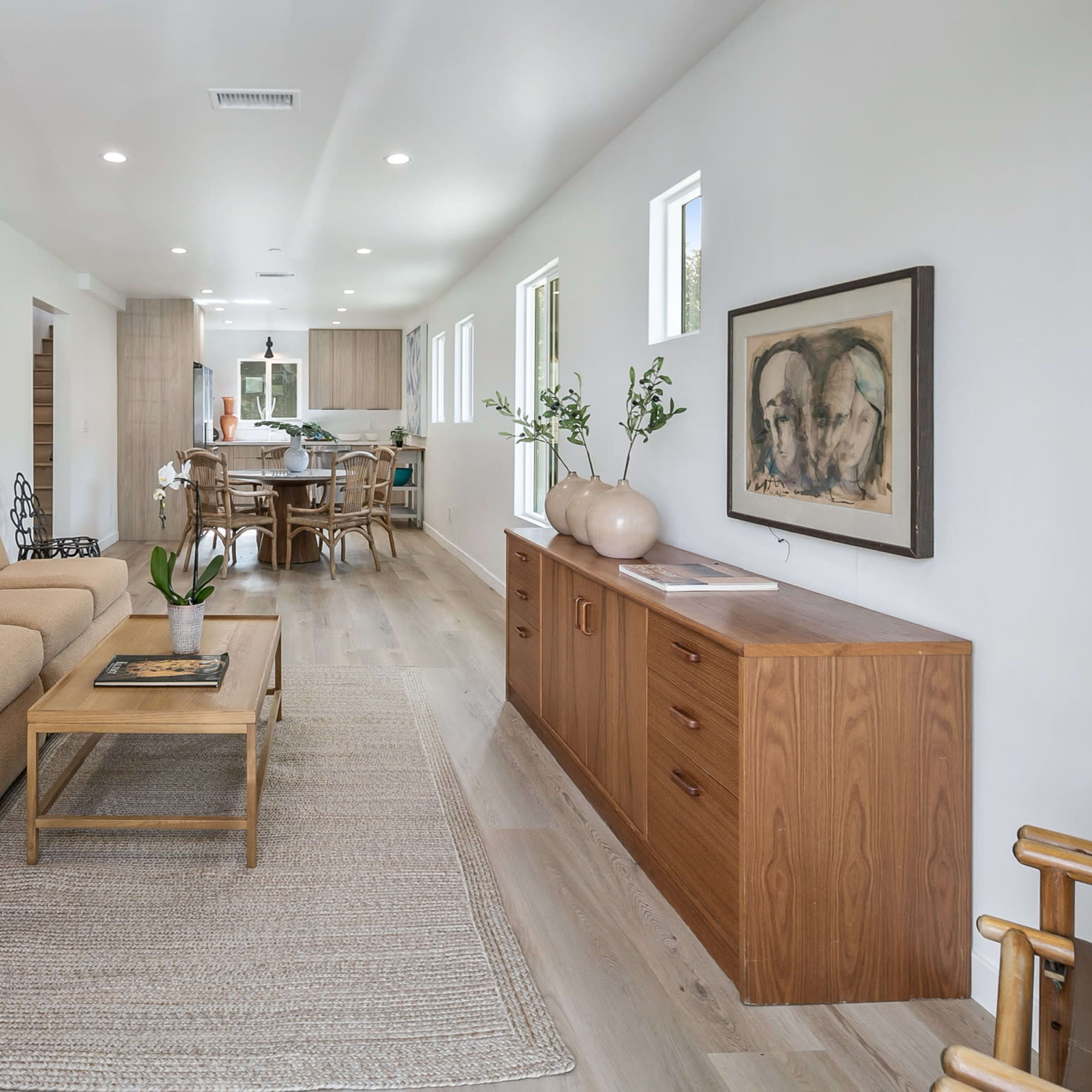 A light-filled living room features a beige sofa, a wooden coffee table, and a sideboard, with a view of an adjoining open kitchen.