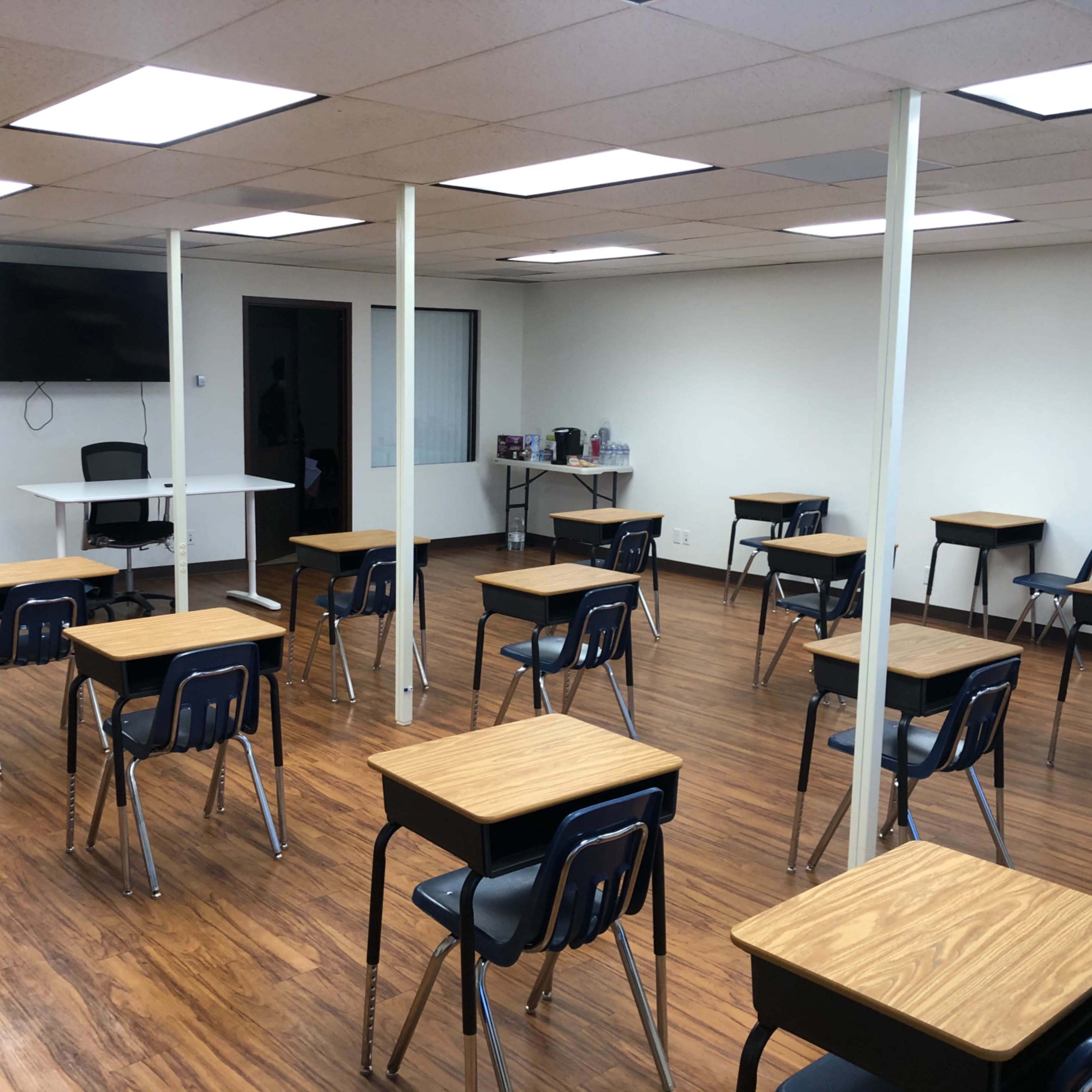 An empty classroom with several desks arranged in rows, a television on the wall, and a small table at the front.