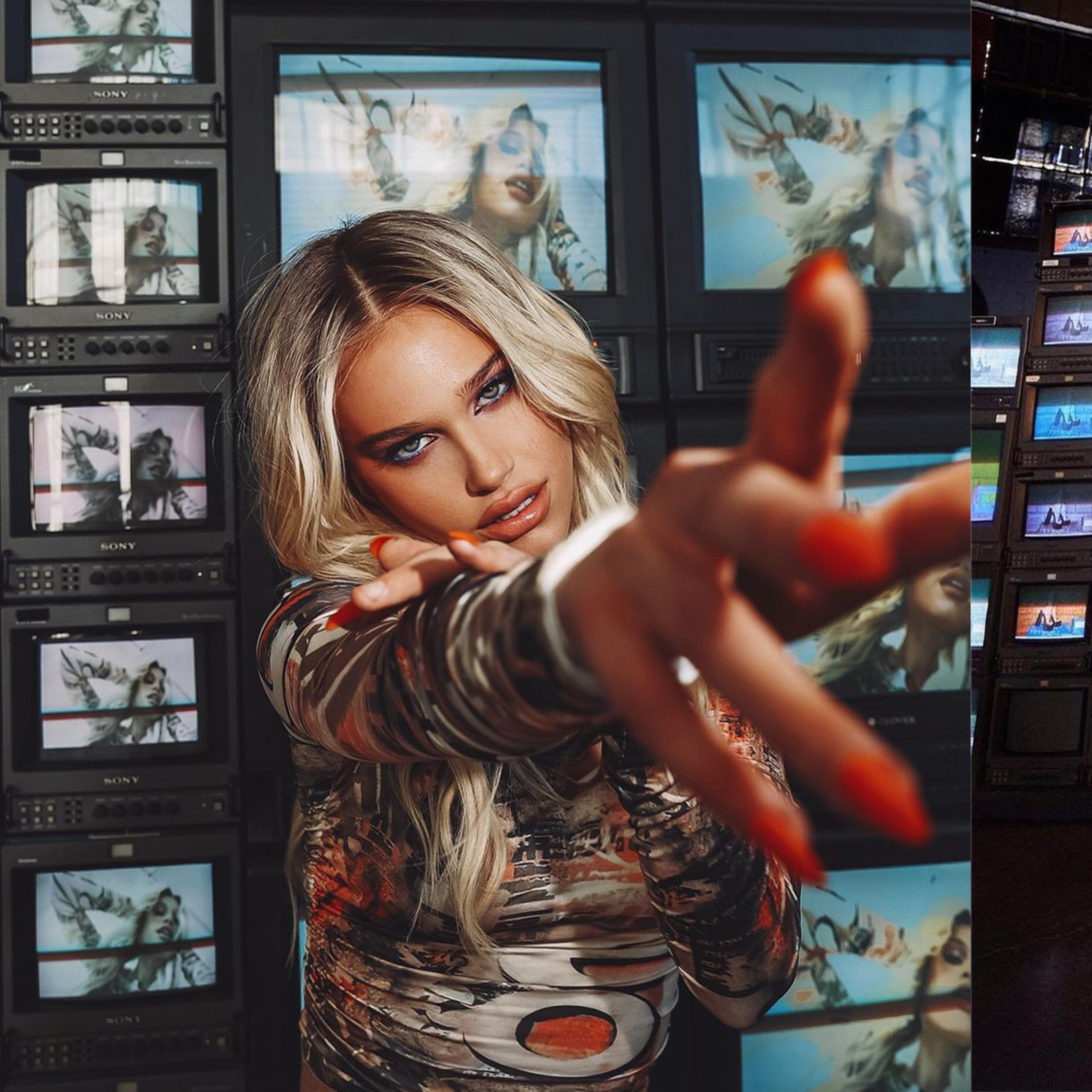 A model poses in front of multiple vintage televisions displaying vibrant images, while reaching out with one hand and wearing a bold outfit.