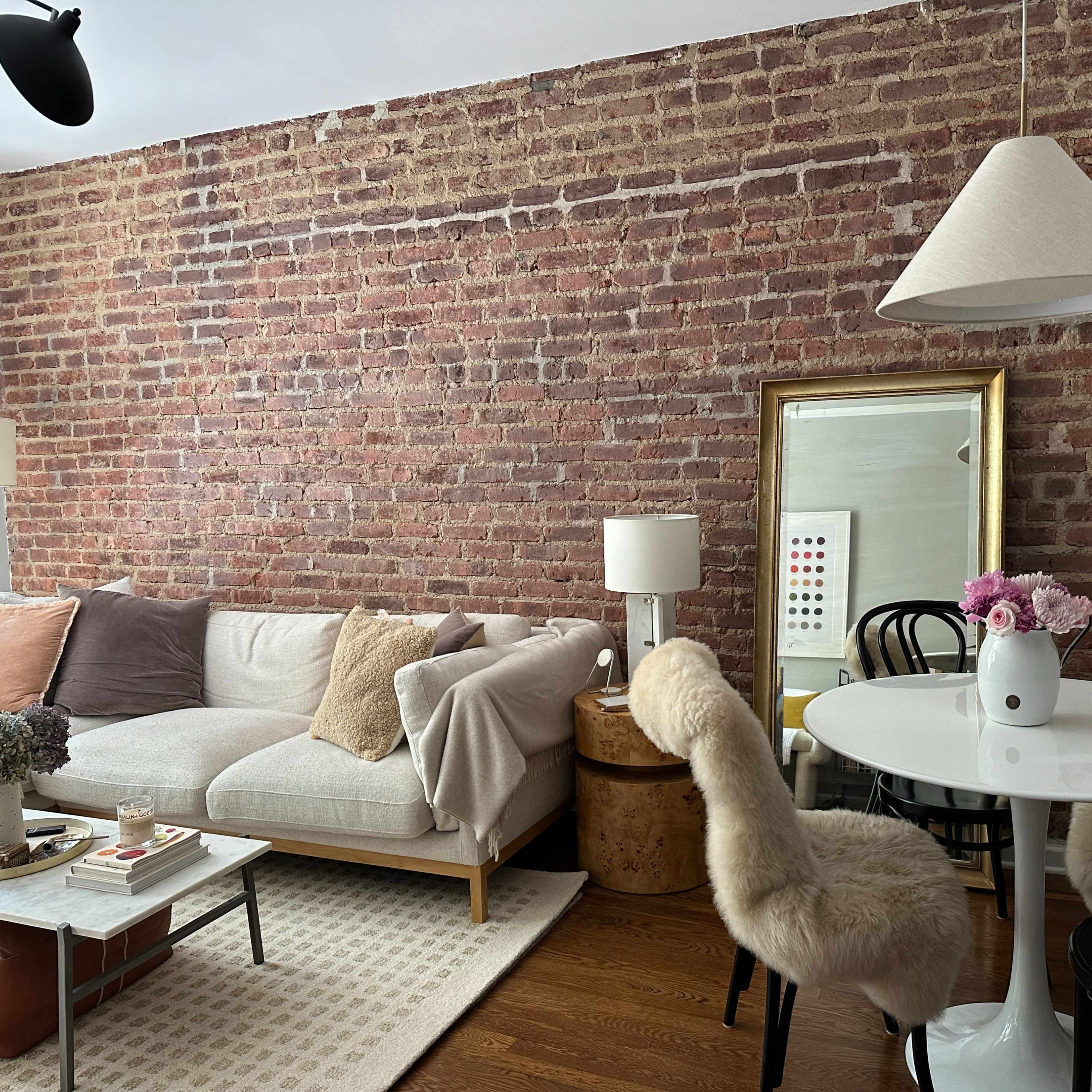 A cozy living space with a brick wall, a neutral sofa, a circular dining table with black chairs, and a decorative mirror.