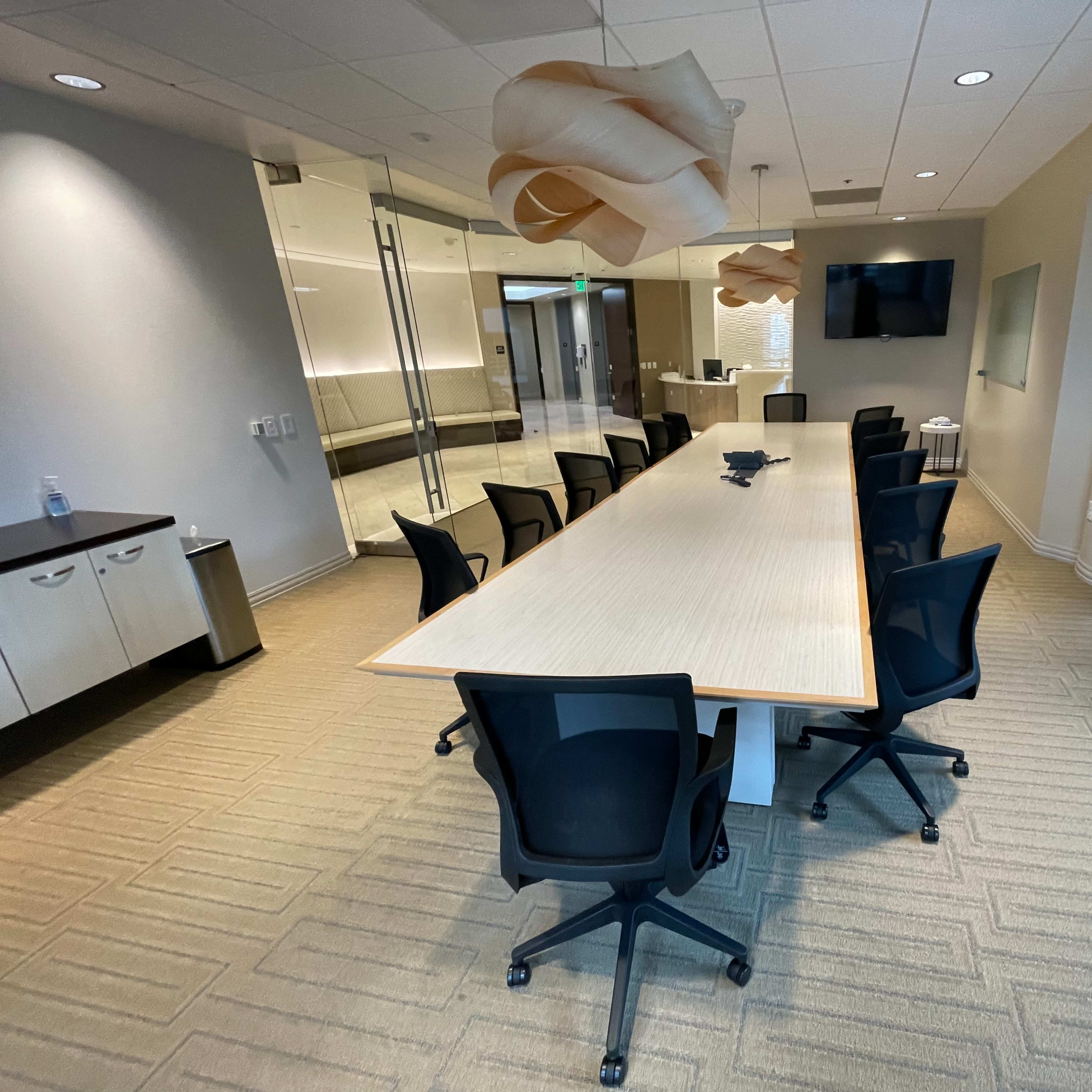 A modern conference room features a long table surrounded by black swivel chairs, with glass walls and overhead lighting.