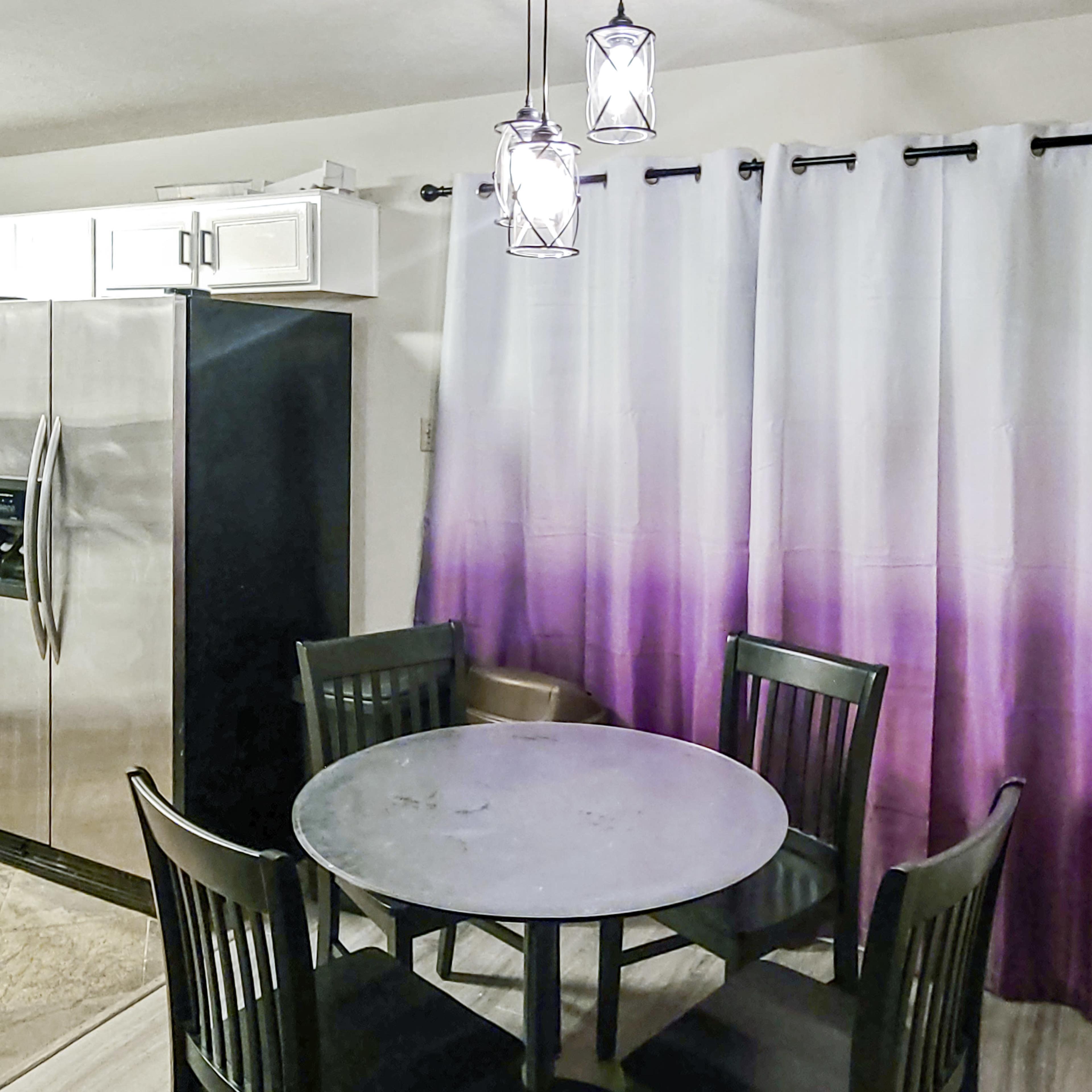A small dining area features a round black table surrounded by four black chairs, with a stainless steel refrigerator and gradient purple curtains in the background.