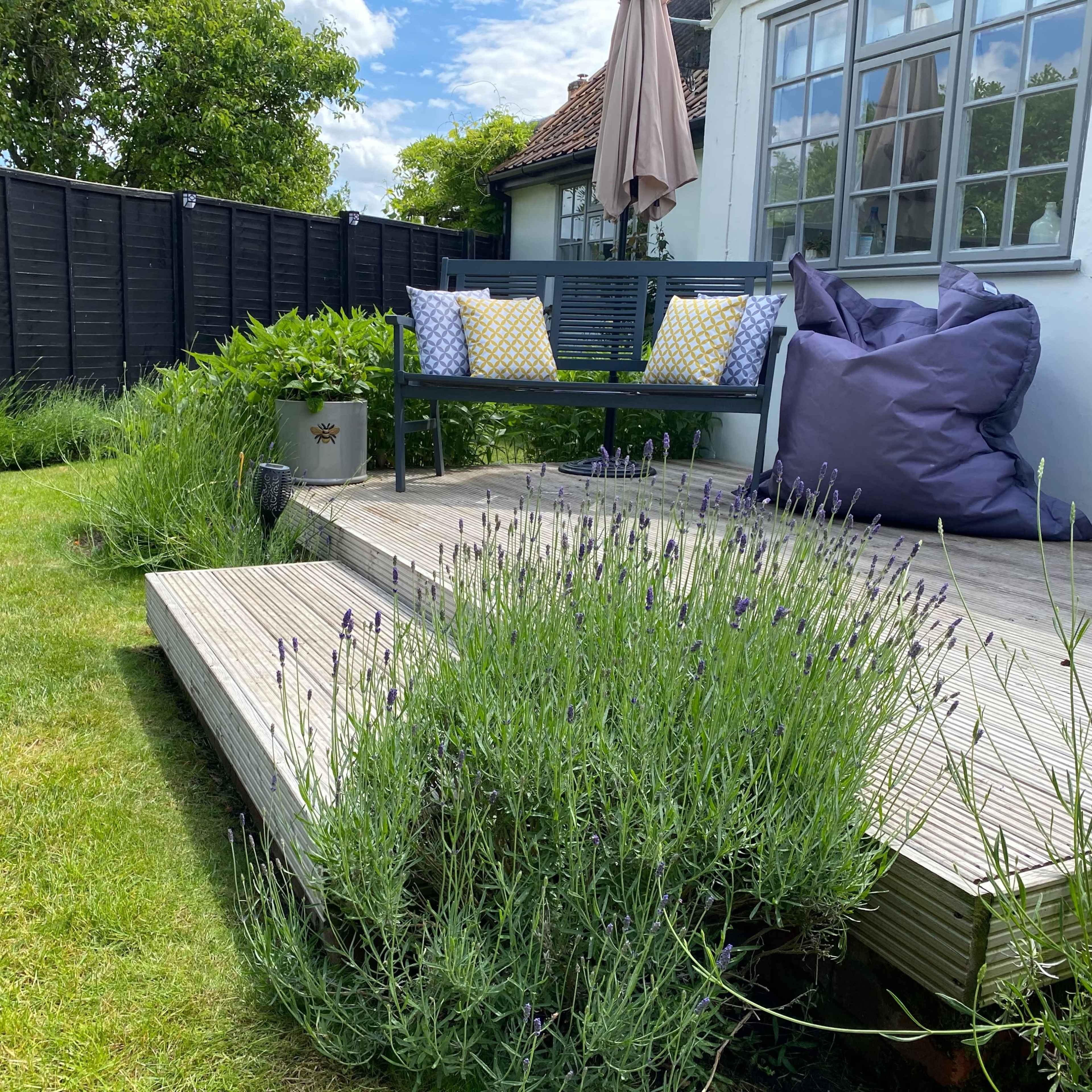 A wooden deck features a sofa with decorative pillows and is surrounded by lavender plants and a grassy area.