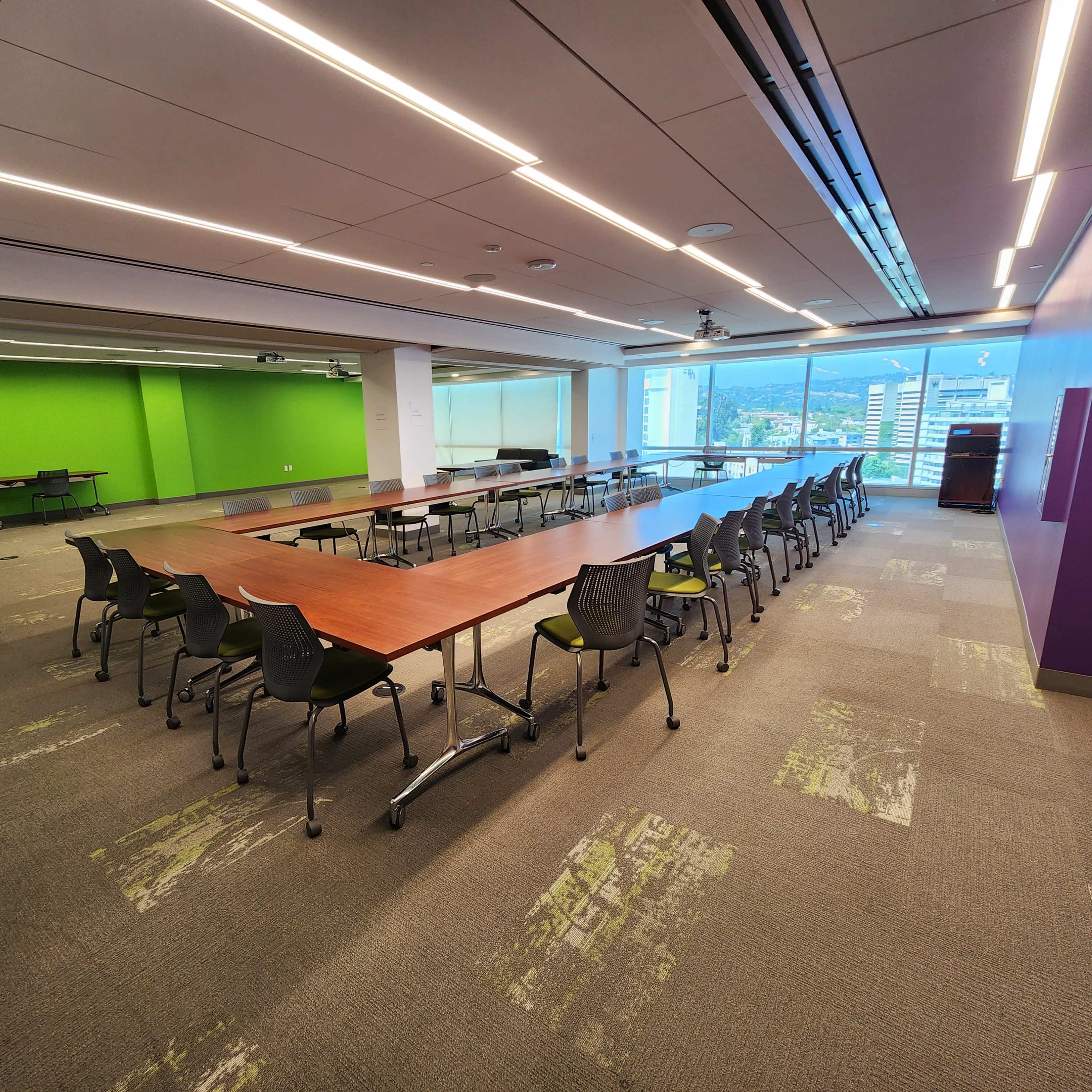 A long conference table is set up in a brightly lit meeting room with green and purple walls and large windows overlooking a cityscape.