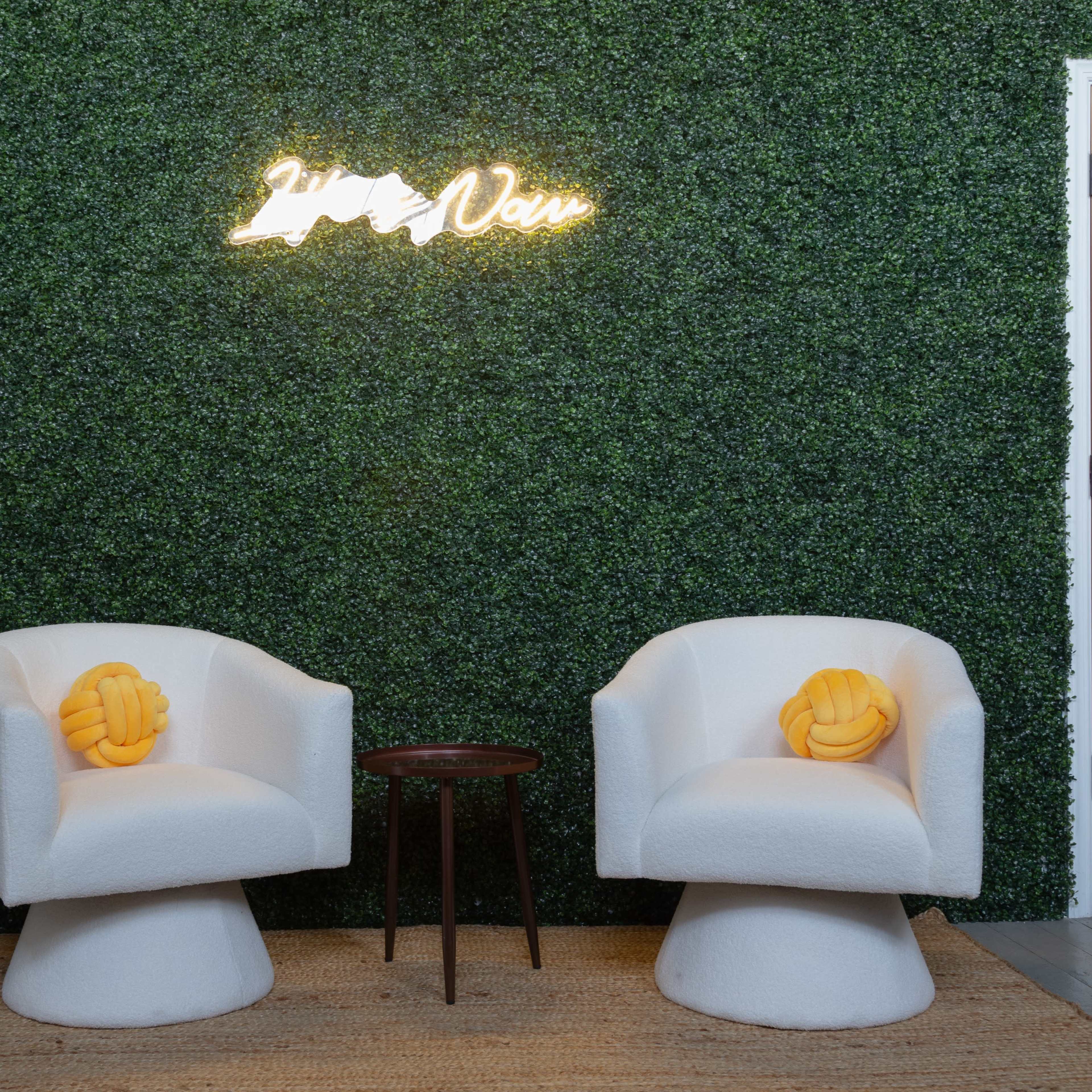 Two white textured chairs with yellow decorative pillows, positioned beside a small brown table against a green foliage wall with a neon sign.