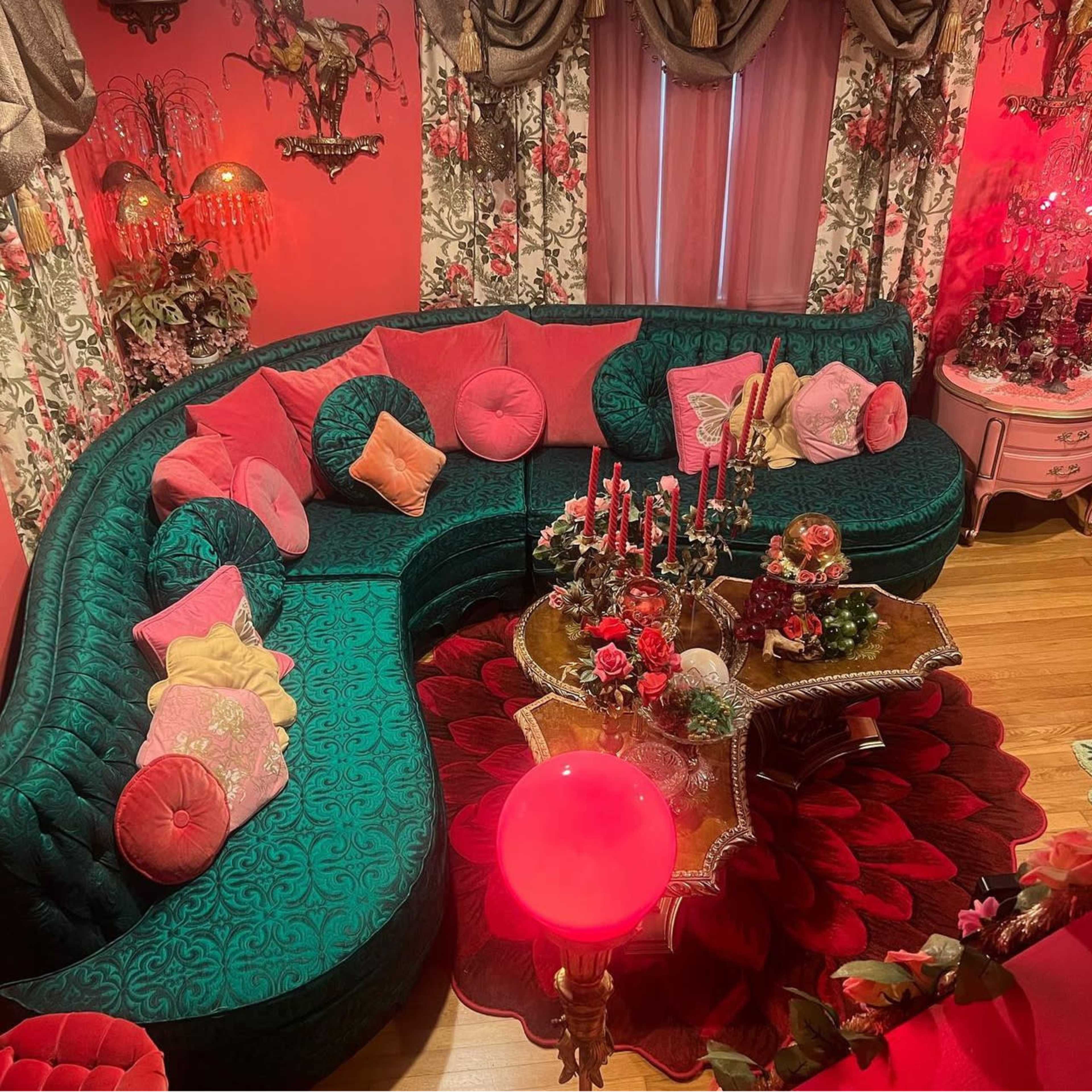 A brightly decorated living room featuring a large green sectional sofa adorned with various cushions, a floral-themed coffee table, and decorative candles, all set against pink walls.