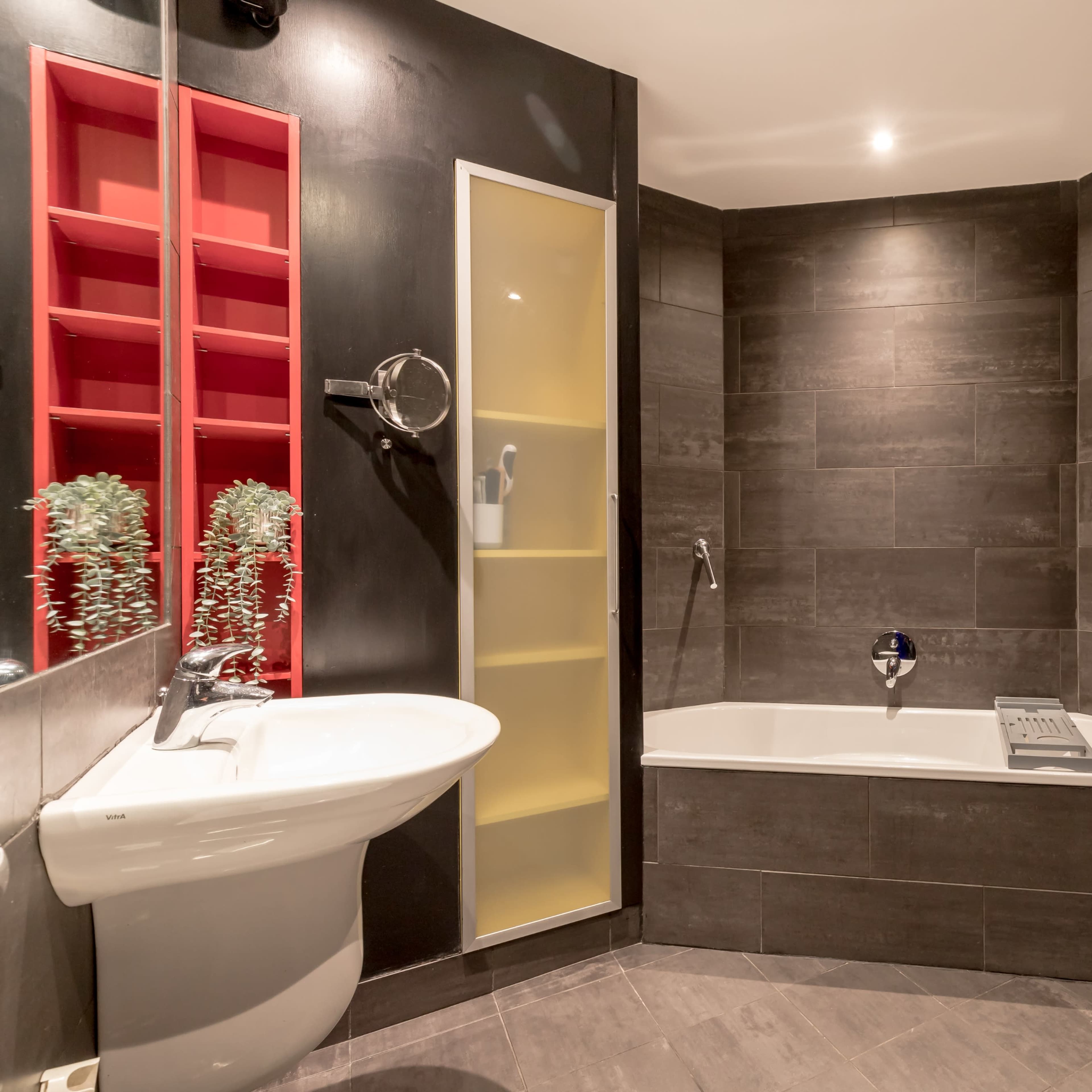 A modern bathroom featuring a white sink, a bathtub, a shower area with a glass door, and a heated towel rack against dark tiled walls.