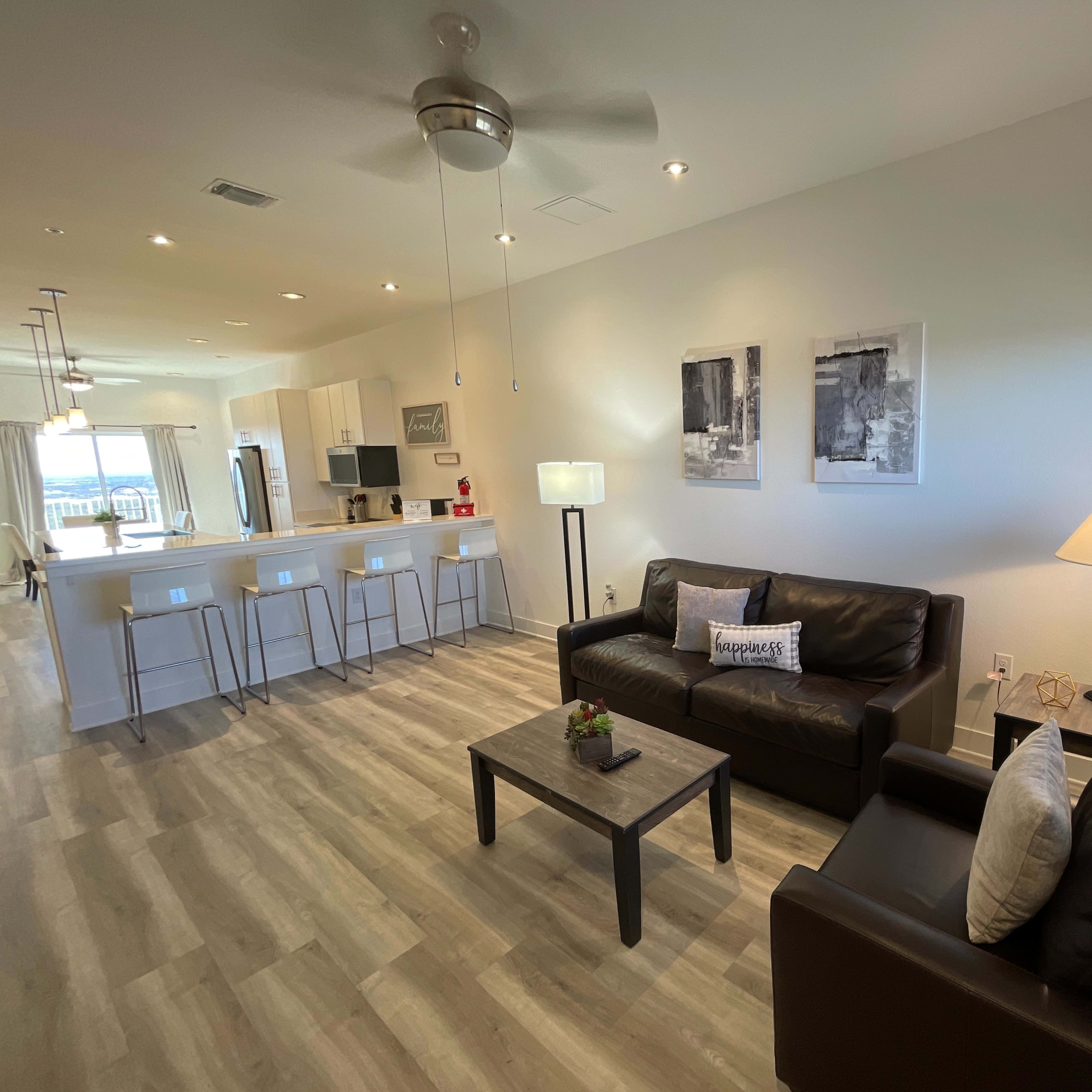 A modern living room with two black leather sofas, a coffee table, and a dining area and kitchen visible in the background.