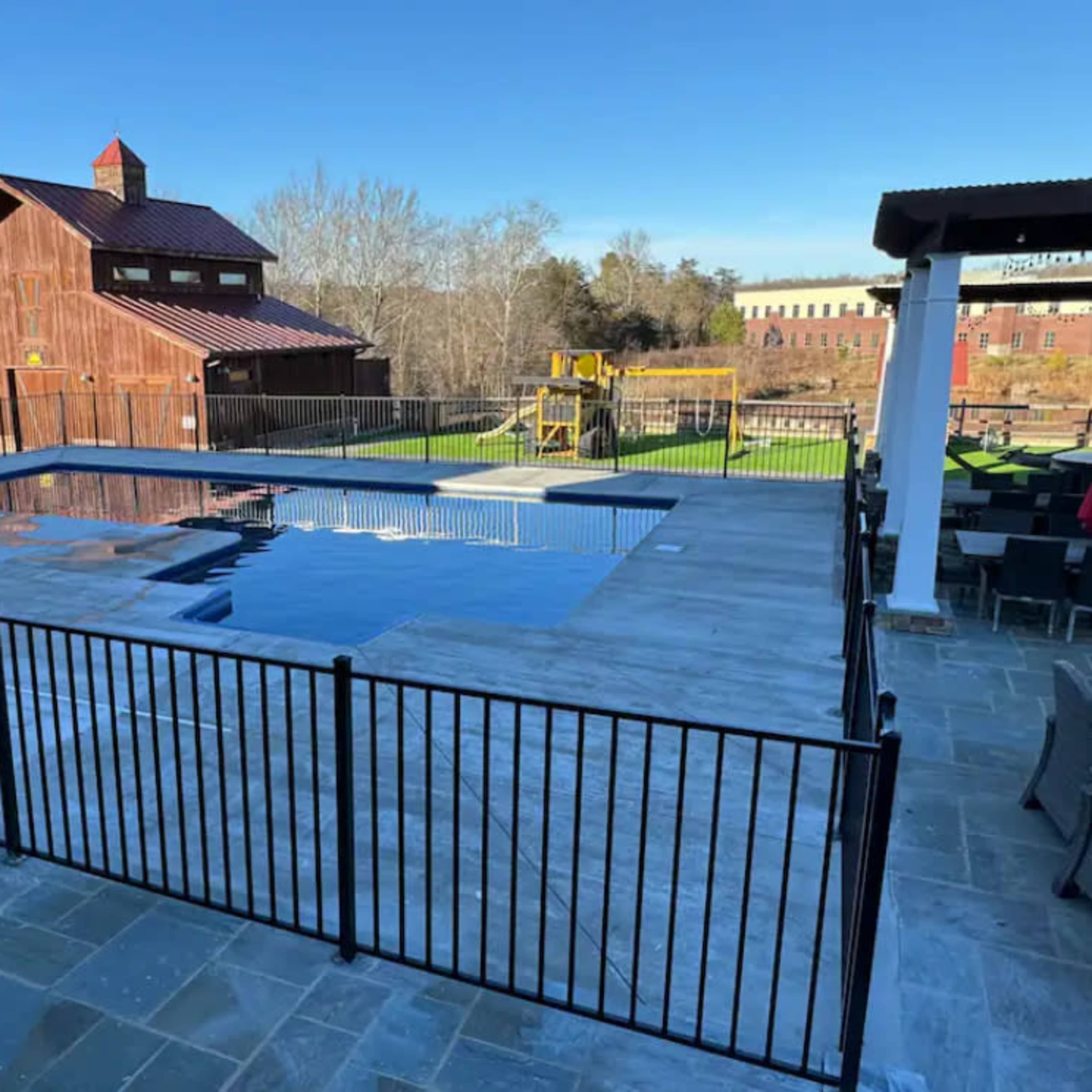 This image shows a swimming pool enclosed by a black fence, with a wooden building and playground equipment visible in the background.