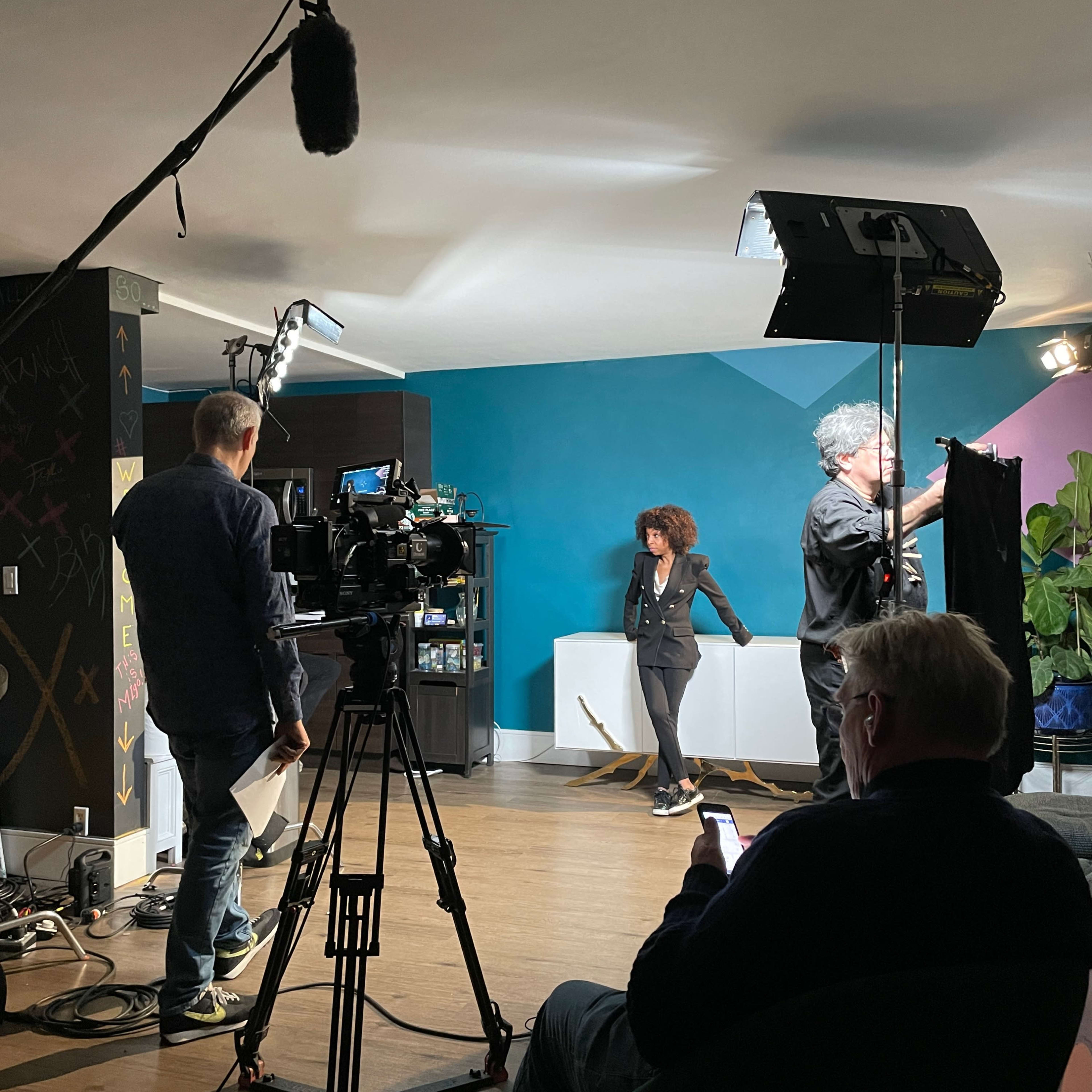 A film crew sets up a shot in a colorful studio with a woman standing in front of a backdrop while a man adjusts lighting equipment.