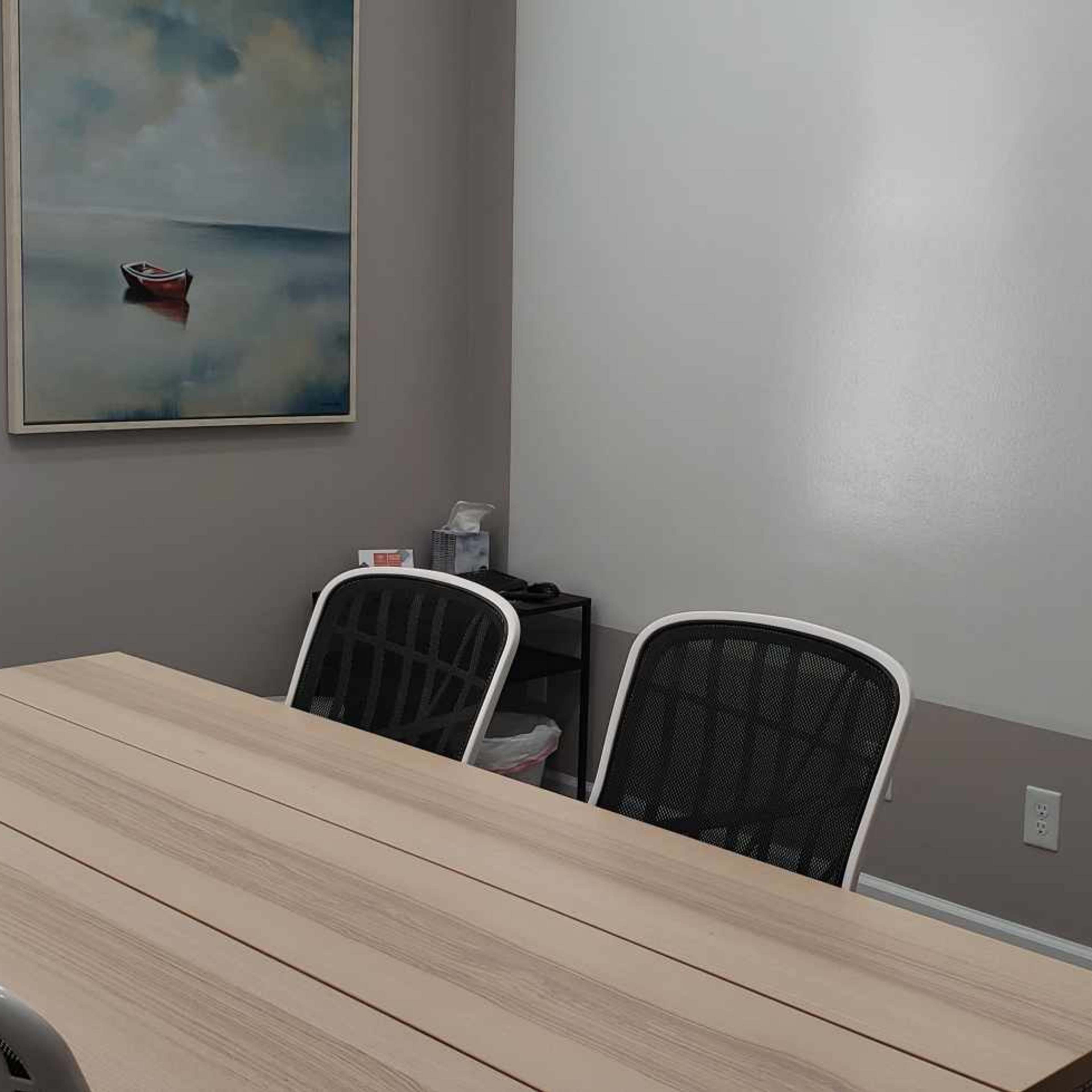 A meeting room features a wooden table surrounded by four chairs, with a painting of a boat on the wall and windows with horizontal blinds.