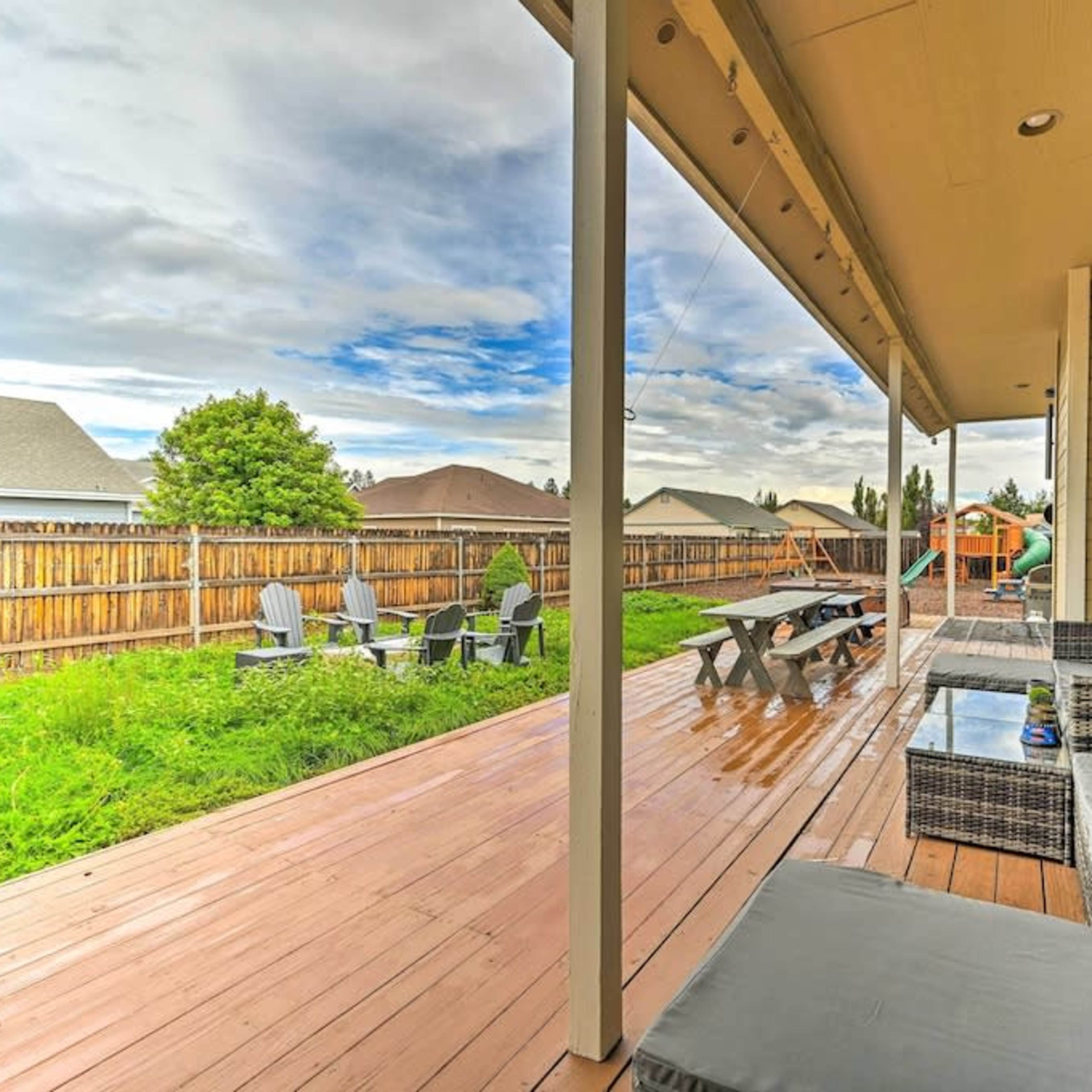 A covered wooden deck features outdoor seating and dining areas, overlooking a grassy yard with a wooden fence in the background.