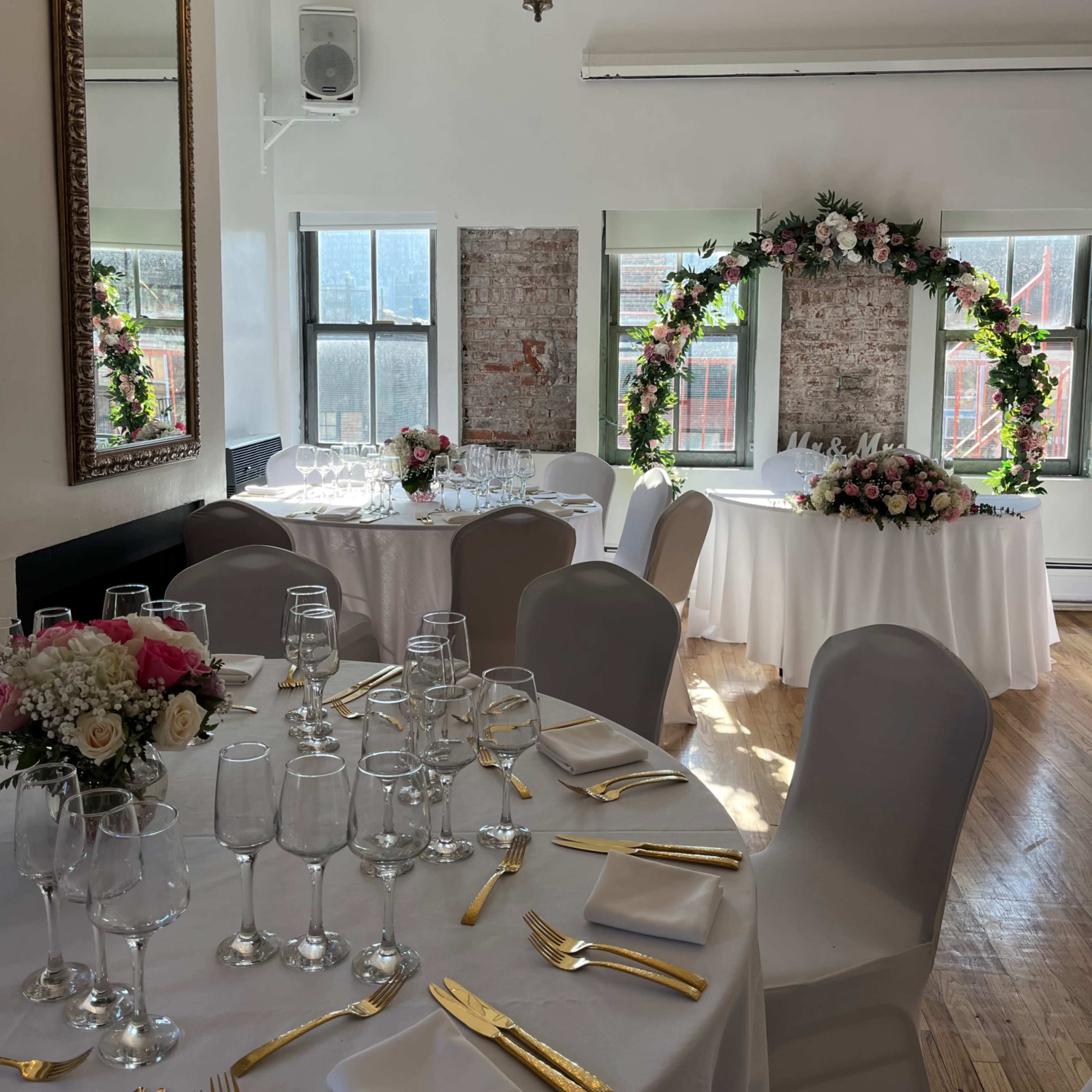 A elegantly decorated event space features a round table adorned with floral arrangements and surrounded by seats, with a chandelier overhead and large windows providing natural light.