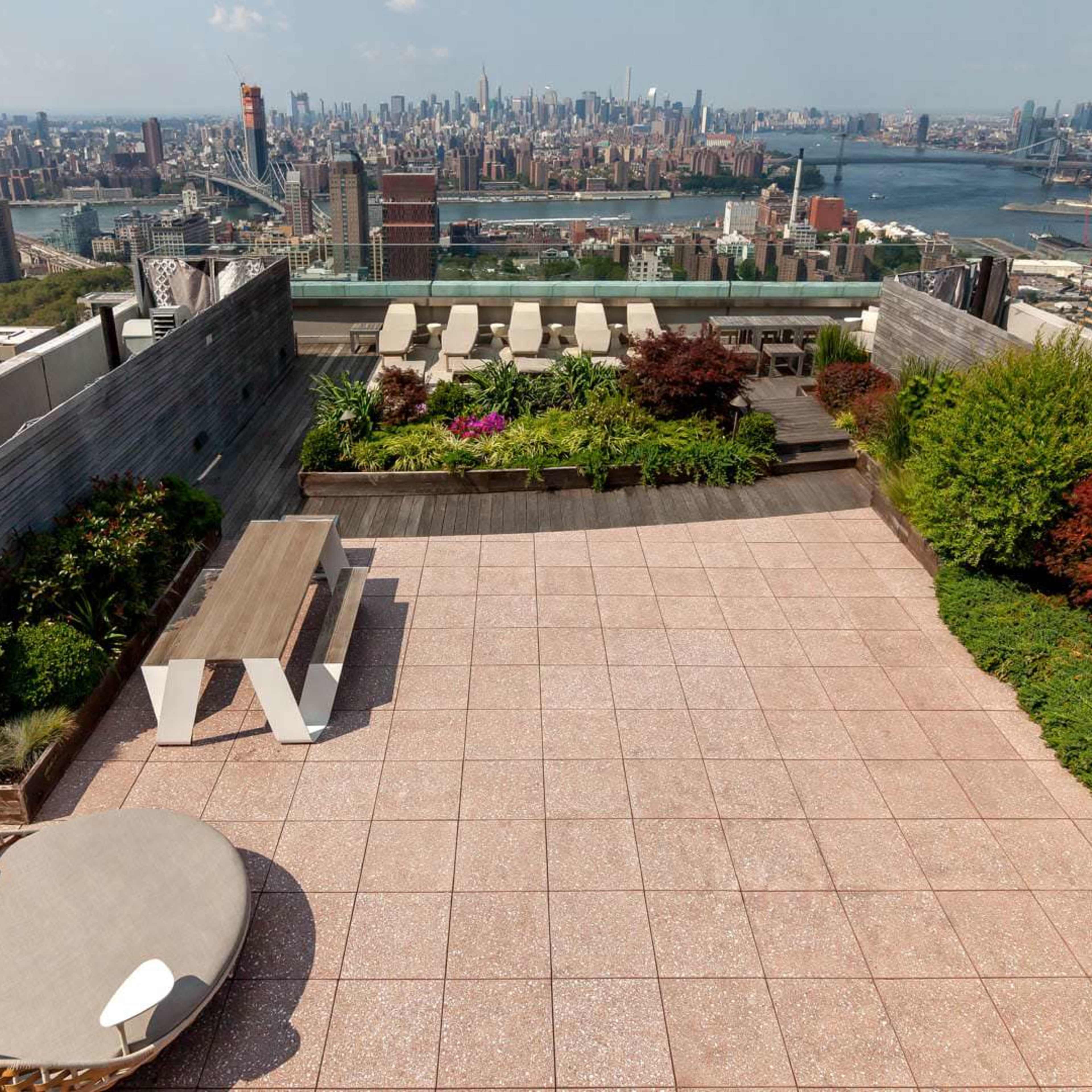 A rooftop terrace with a view of a city skyline, featuring landscaped greenery and outdoor furniture arranged on a tiled surface.