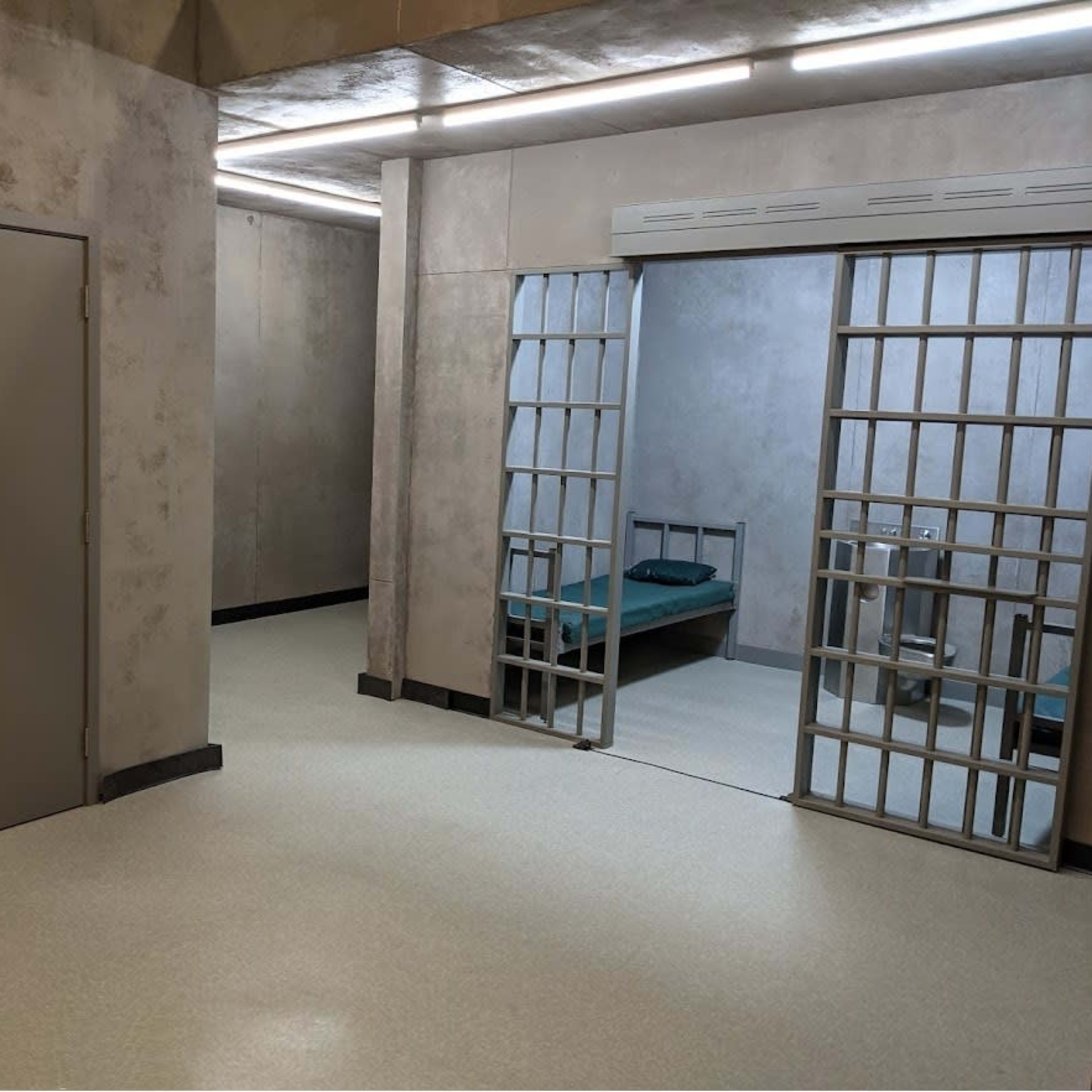 A stark, industrial room with two metal doors and a jail cell containing a bed.