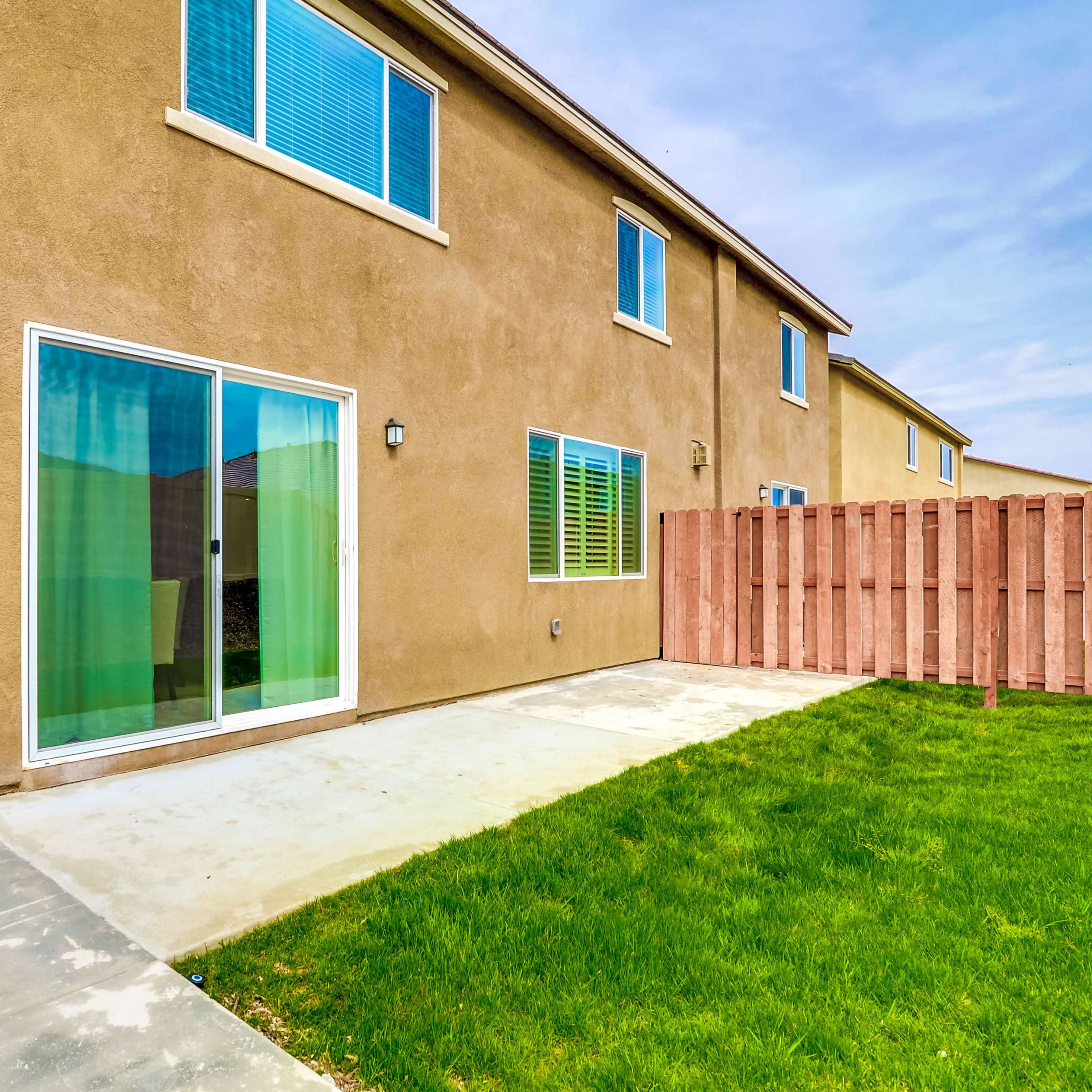 The exterior of a two-story house with a small concrete patio, green grass area, and a wooden fence.