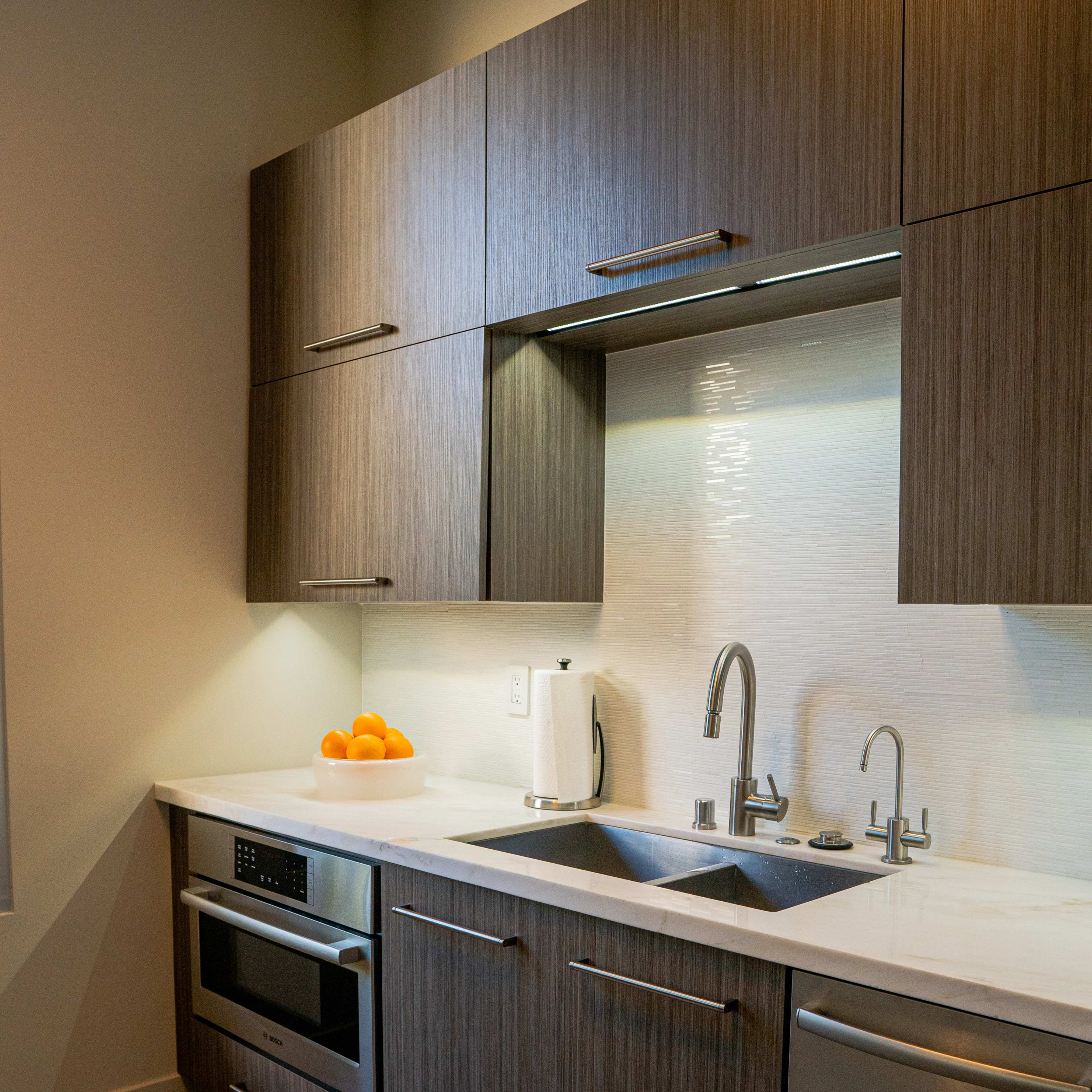 A modern kitchen features wooden cabinets, stainless steel appliances, and a sink with a countertop, complemented by a kettle and a bowl of oranges.