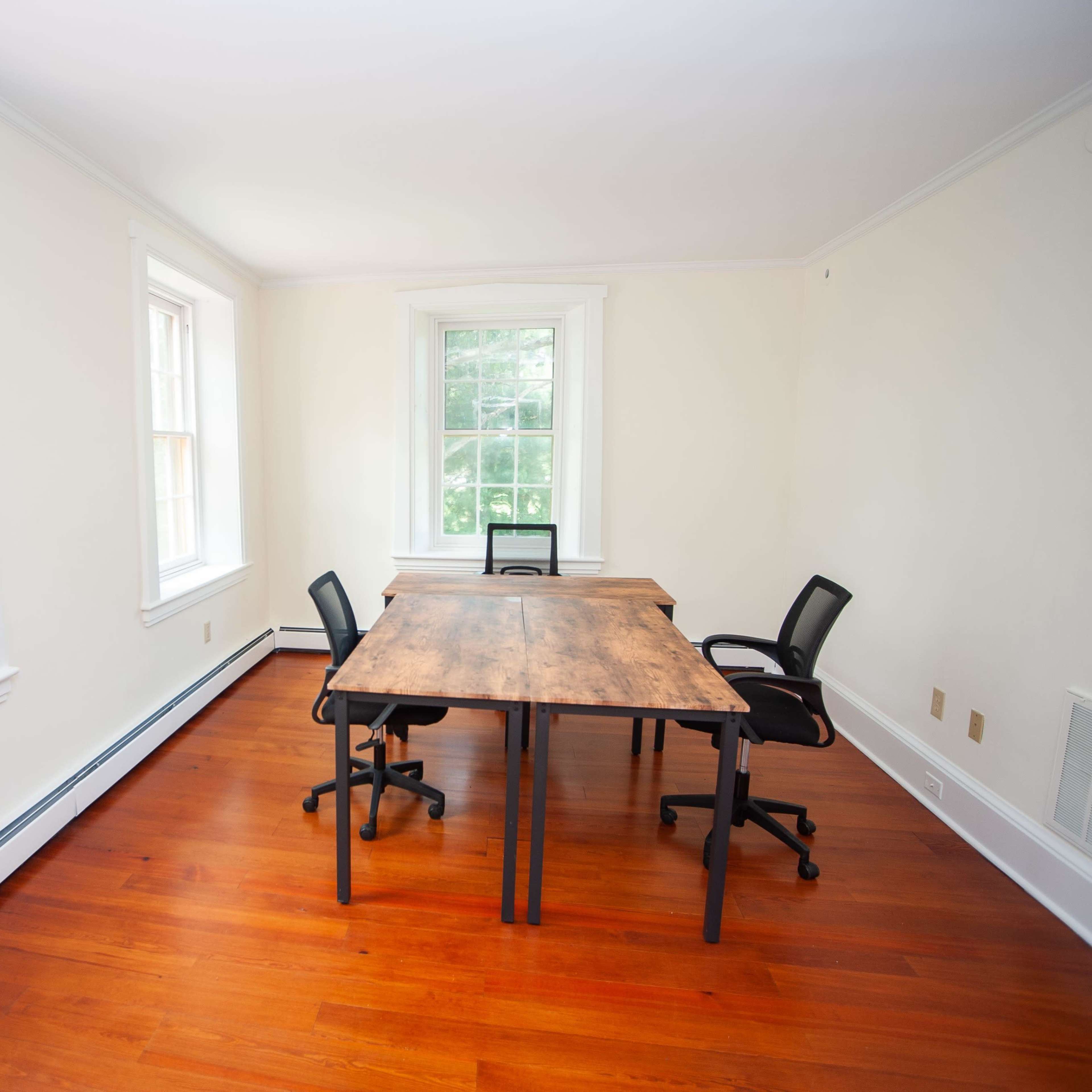 A bright, uncluttered room with two windows, a wooden table, and two black office chairs.